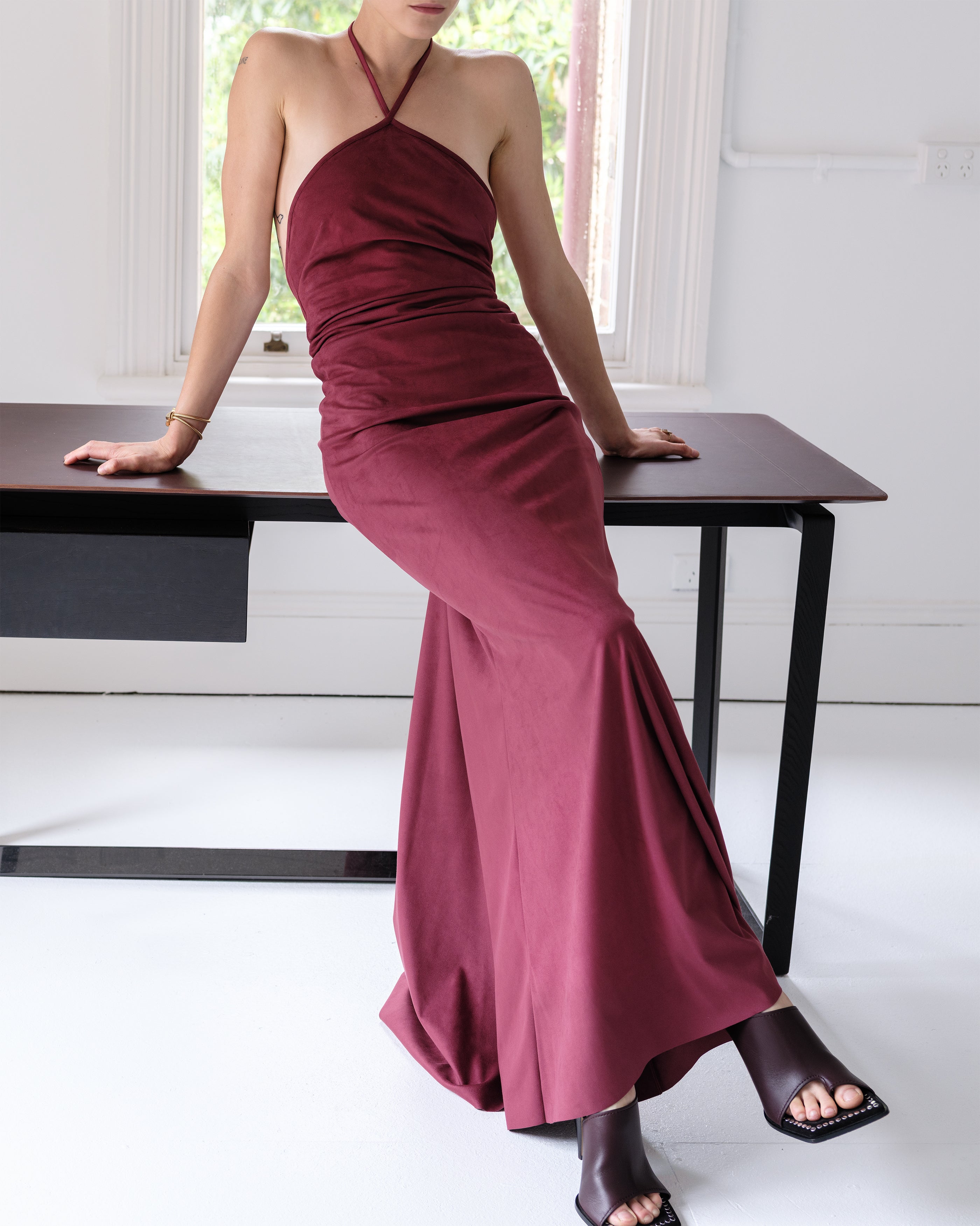 Woman in a burgundy halter neck Common Hours dress sitting on a table indoors.