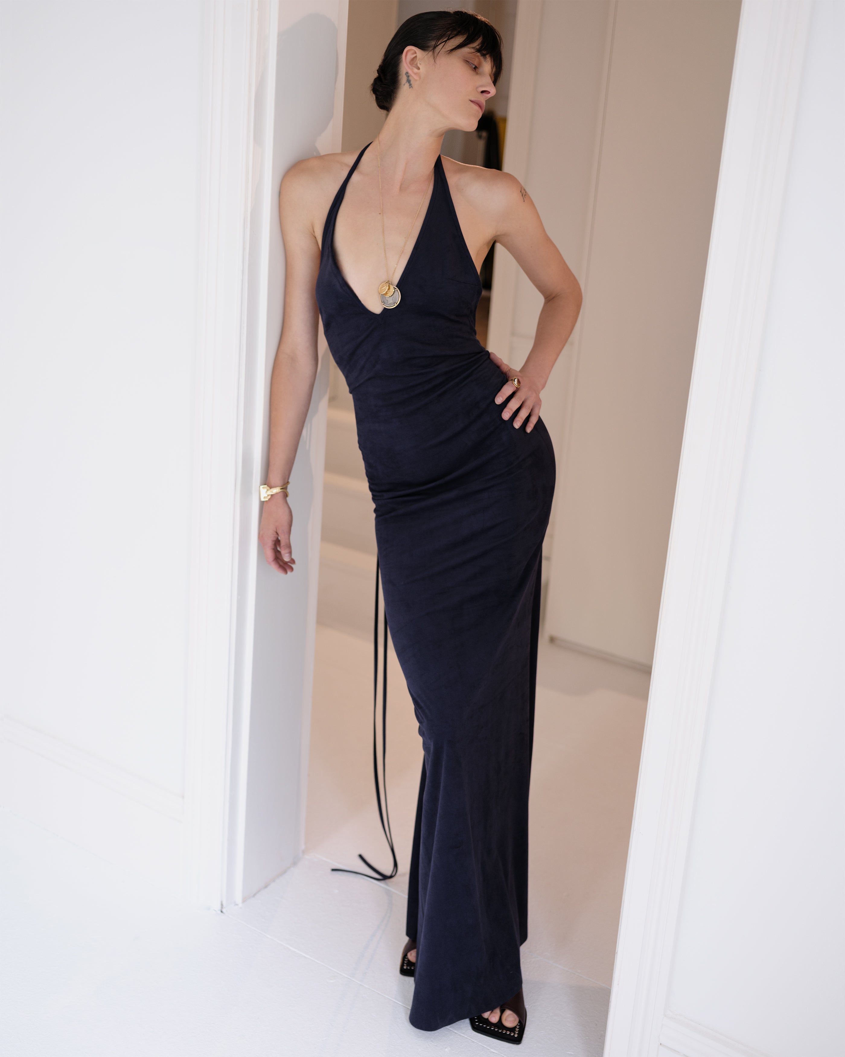 Woman wearing a navy suede Common Hours evening gown with a deep V neckline standing in a doorway.