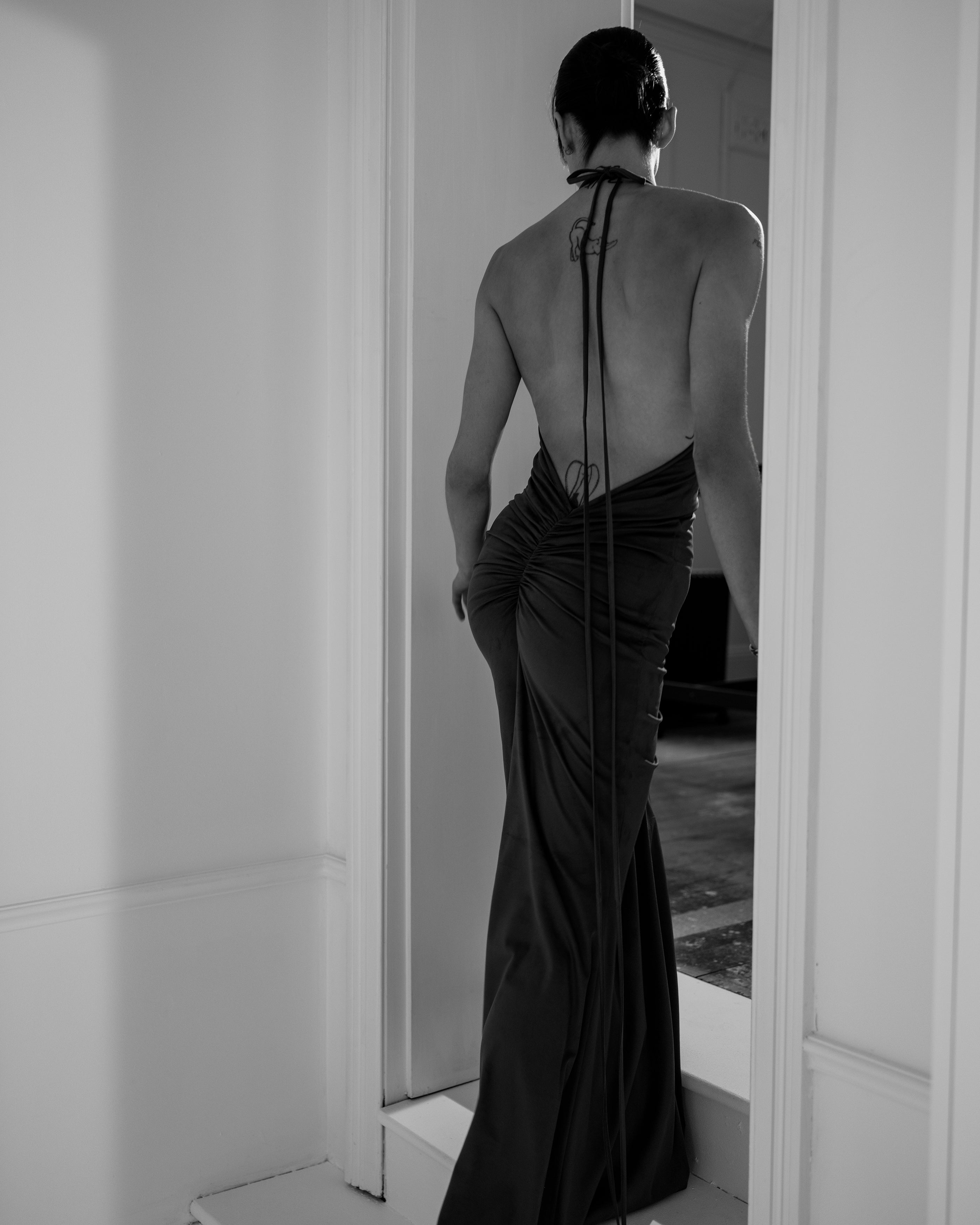 Woman in a long, elegant suede Common Hours dress with an open back standing in a room.