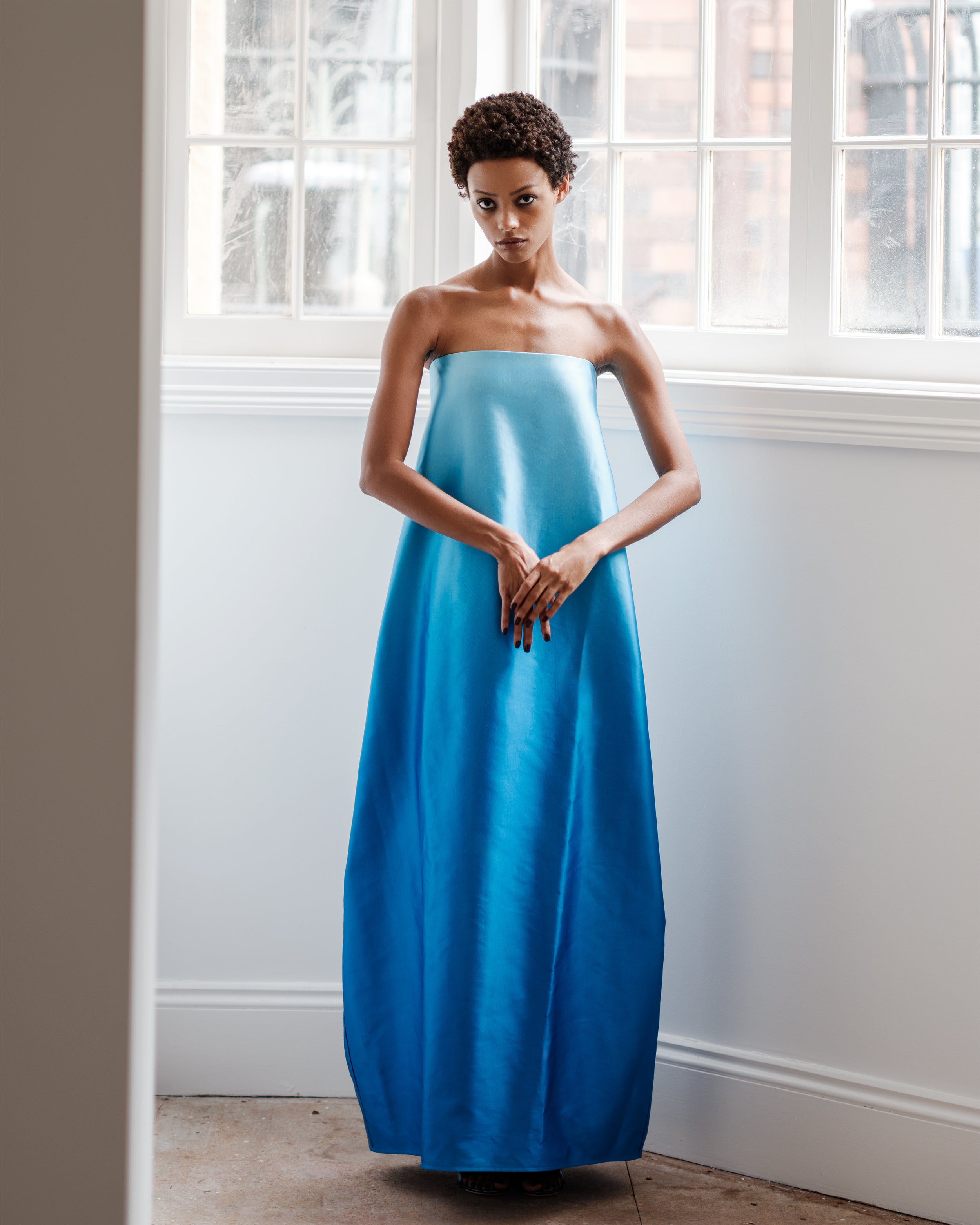 Woman wearing a strapless blue maxi dress Entourage by Common Hours with large windows in background.