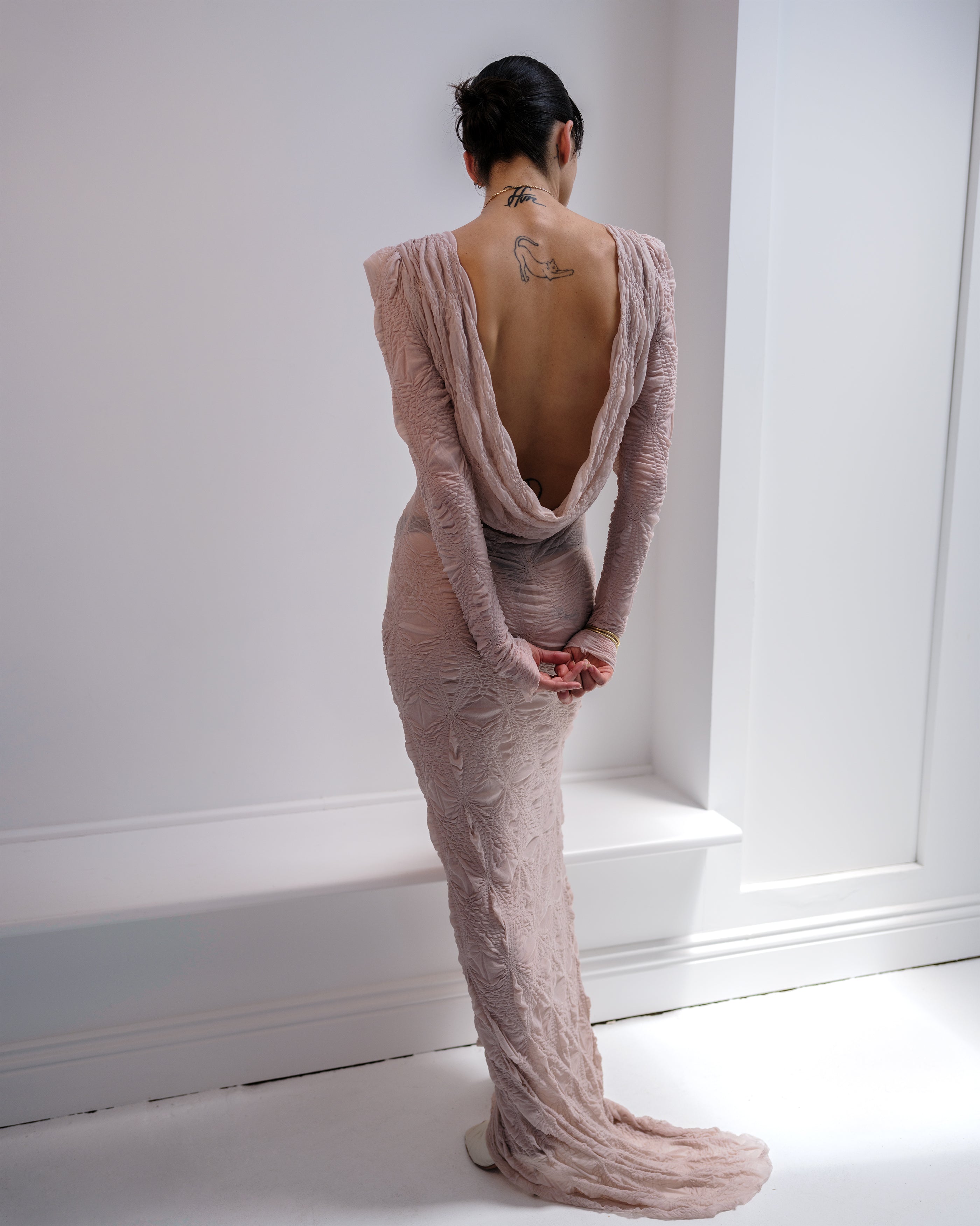 Woman wearing a pink textured Intention Common Hours dress with a backless design, standing against a white wall.