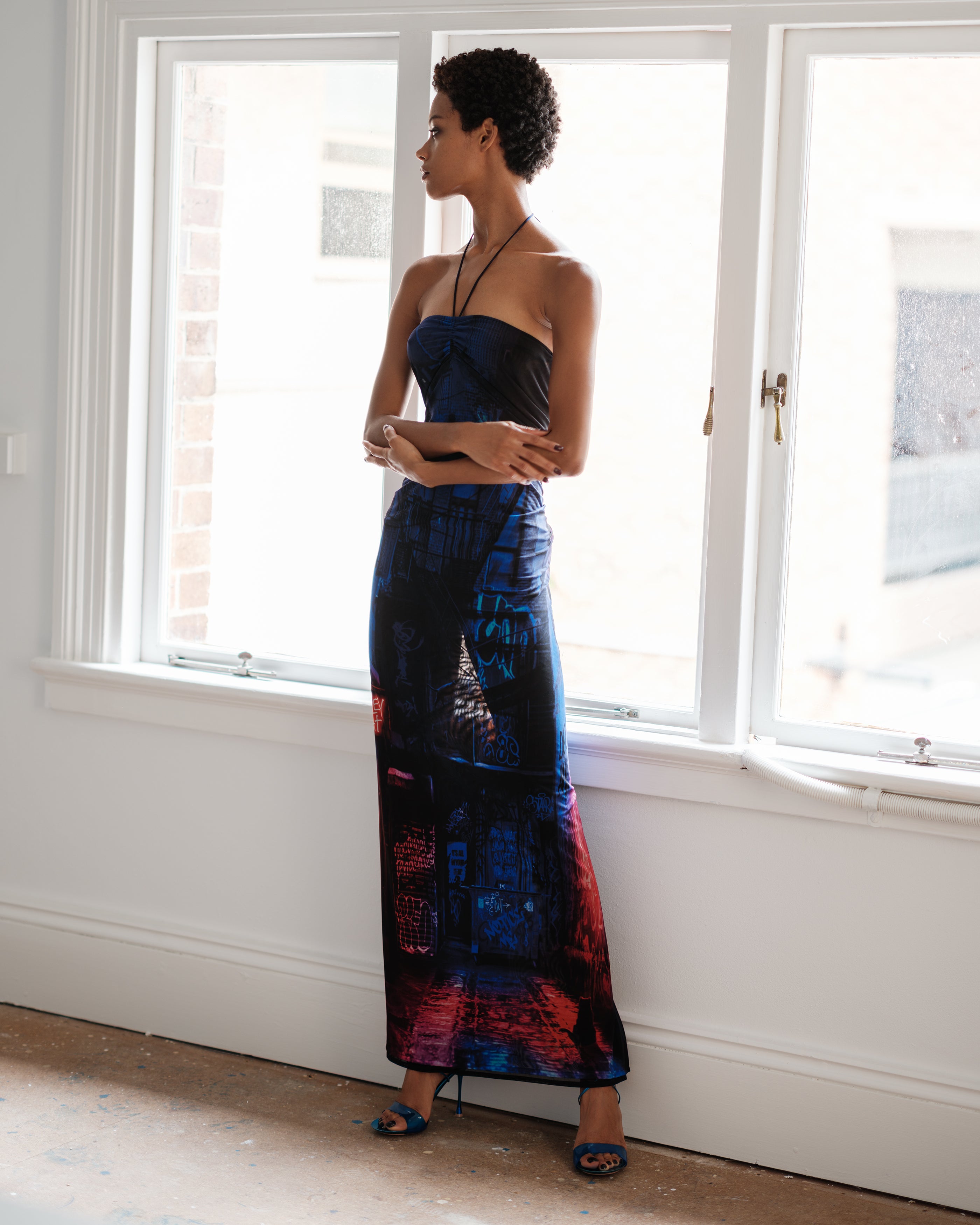 Woman in a long, dark blue Common Hours Just Us dress with abstract alleycat and graffiti patterns standing in a room with large windows.