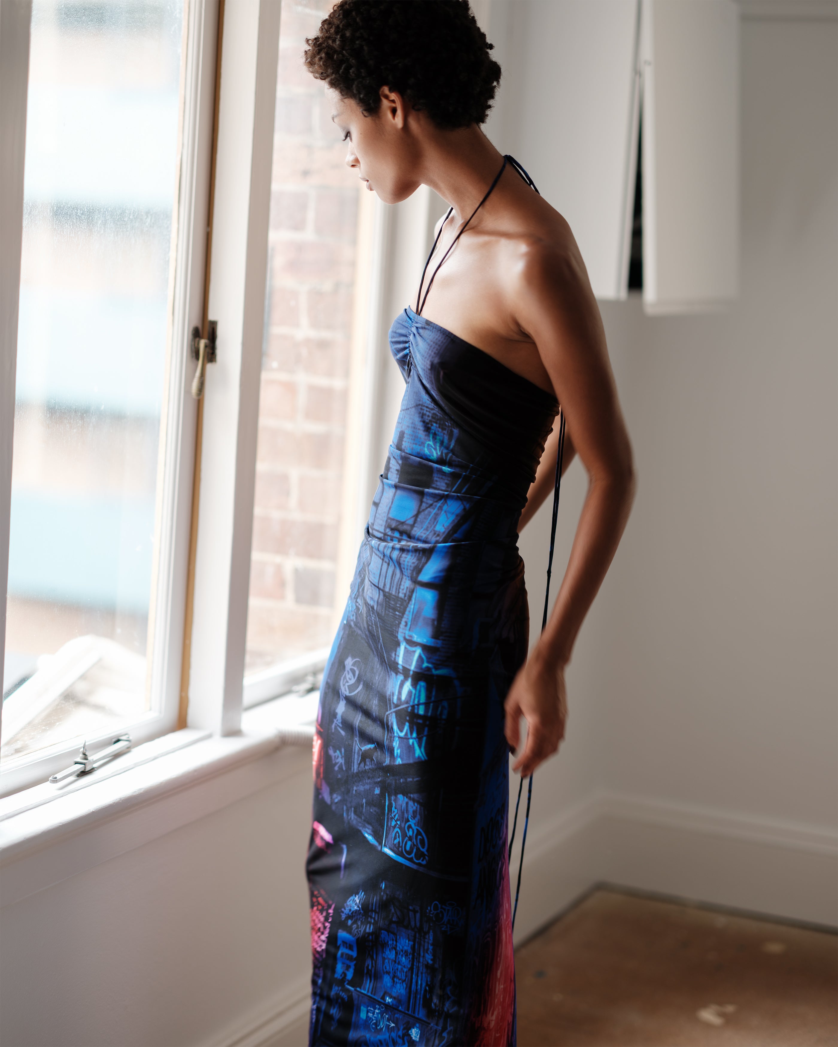 Woman in a Common Hours Just Us halter neck dress standing by a window.