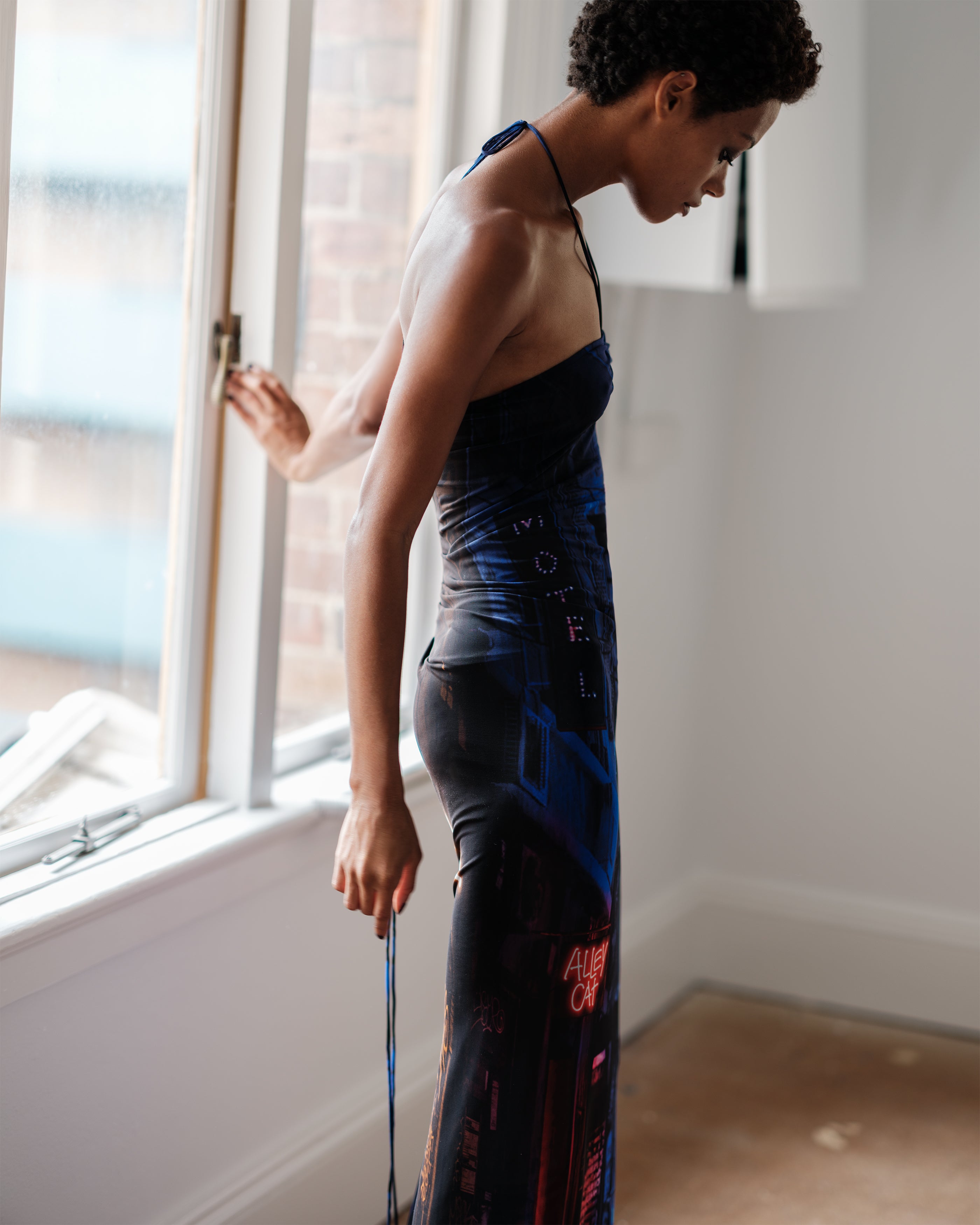 Woman in a halter neck Common Hours Just Us dress standing by a window.