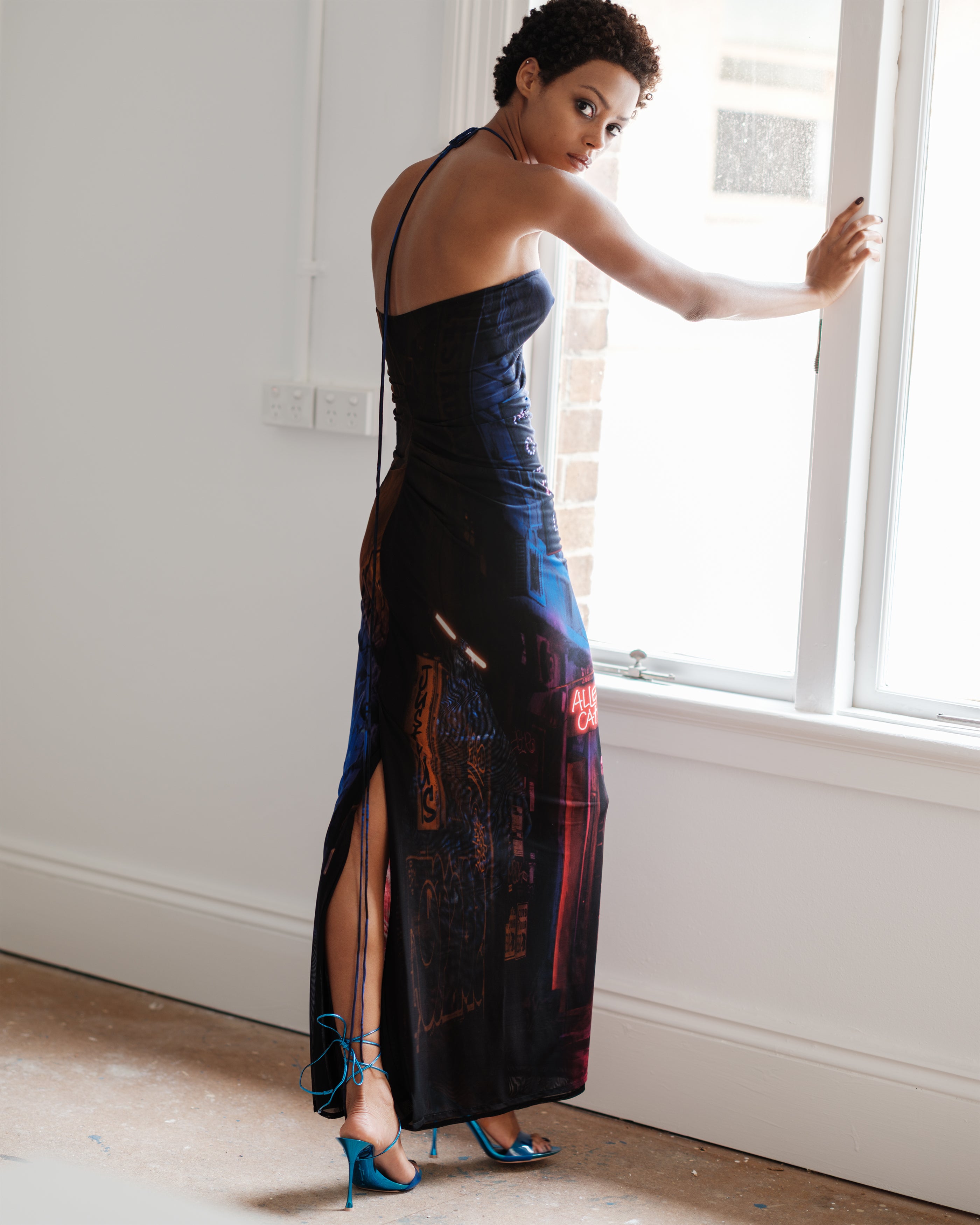 Woman in a Common Hours Just Us evening gown with a high slit standing by a window.