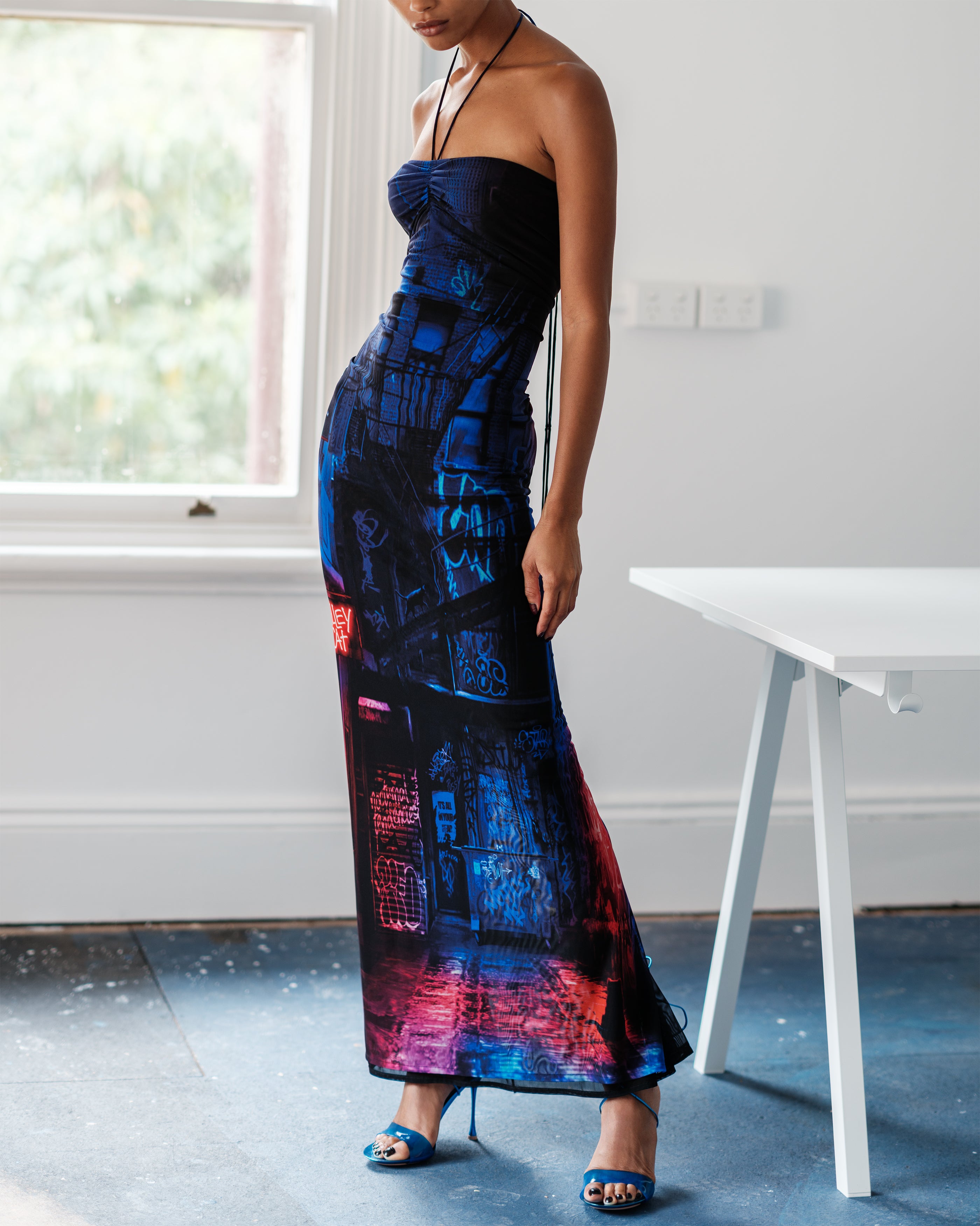 Woman wearing a halter neck Common Hours Just Us dress with a colorful abstract pattern in a room with a window and table.