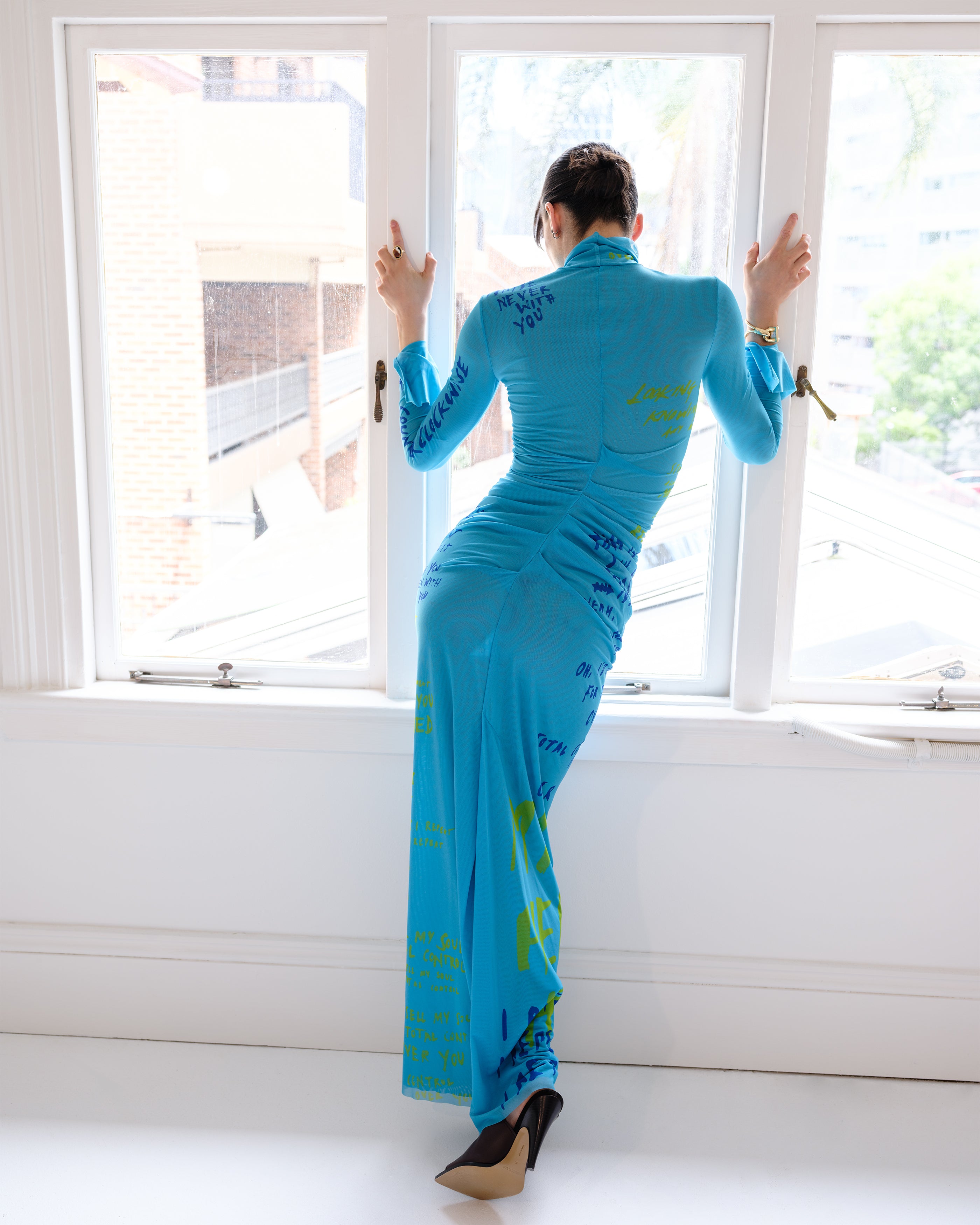 Woman wearing a Common Hours Motels blue dress with text patterns standing in front of a window.
