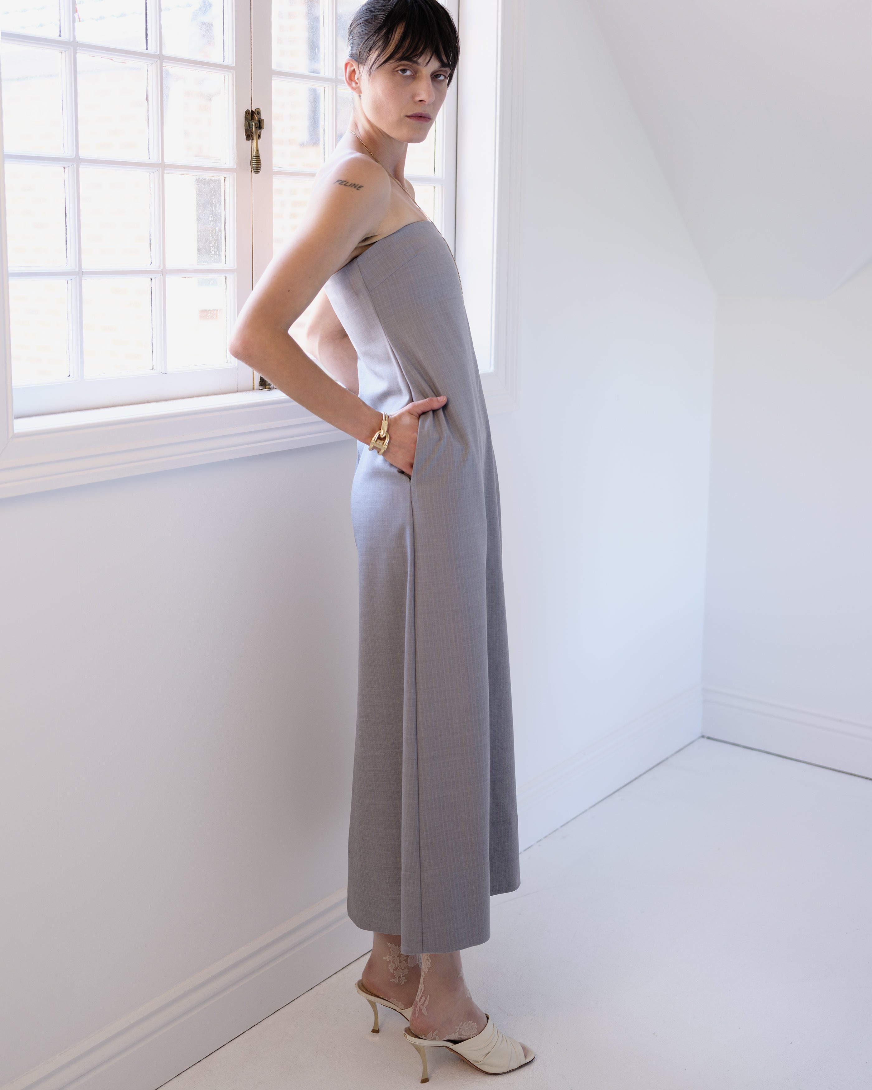 Woman in a strapless gray Common Hours tailored dress standing in a bright room with white walls and a window.