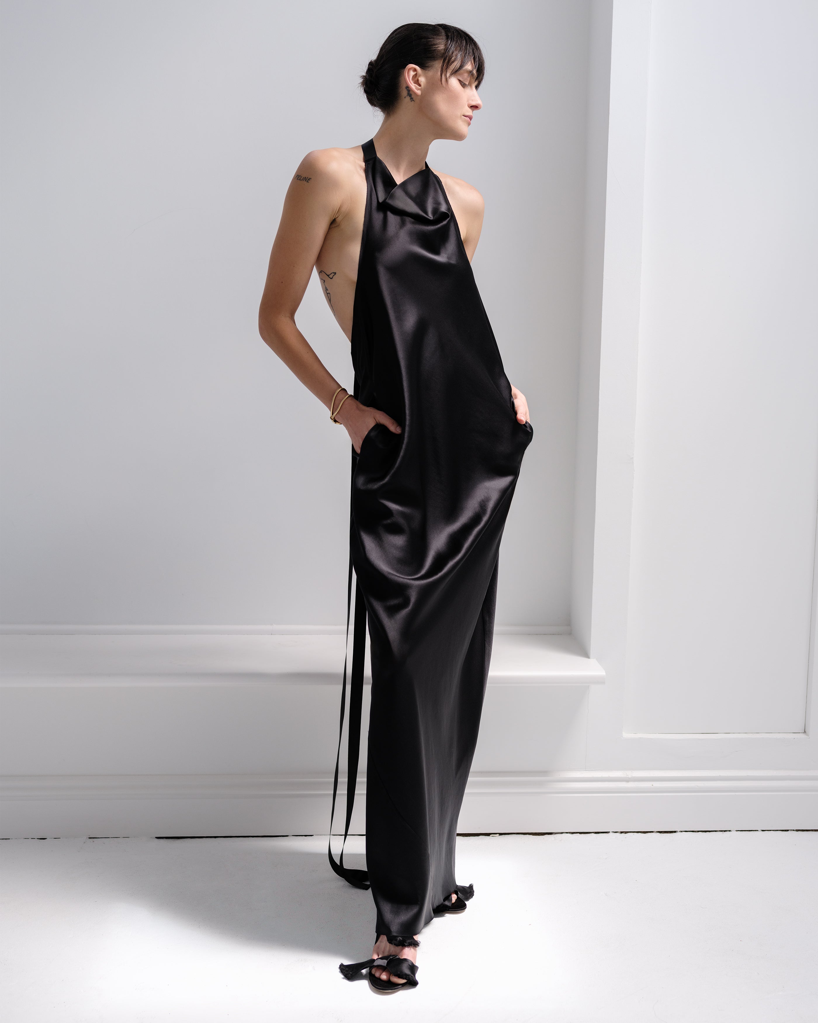 Woman wearing a black satin Common Hours dress with ribbon ties standing against a white wall.