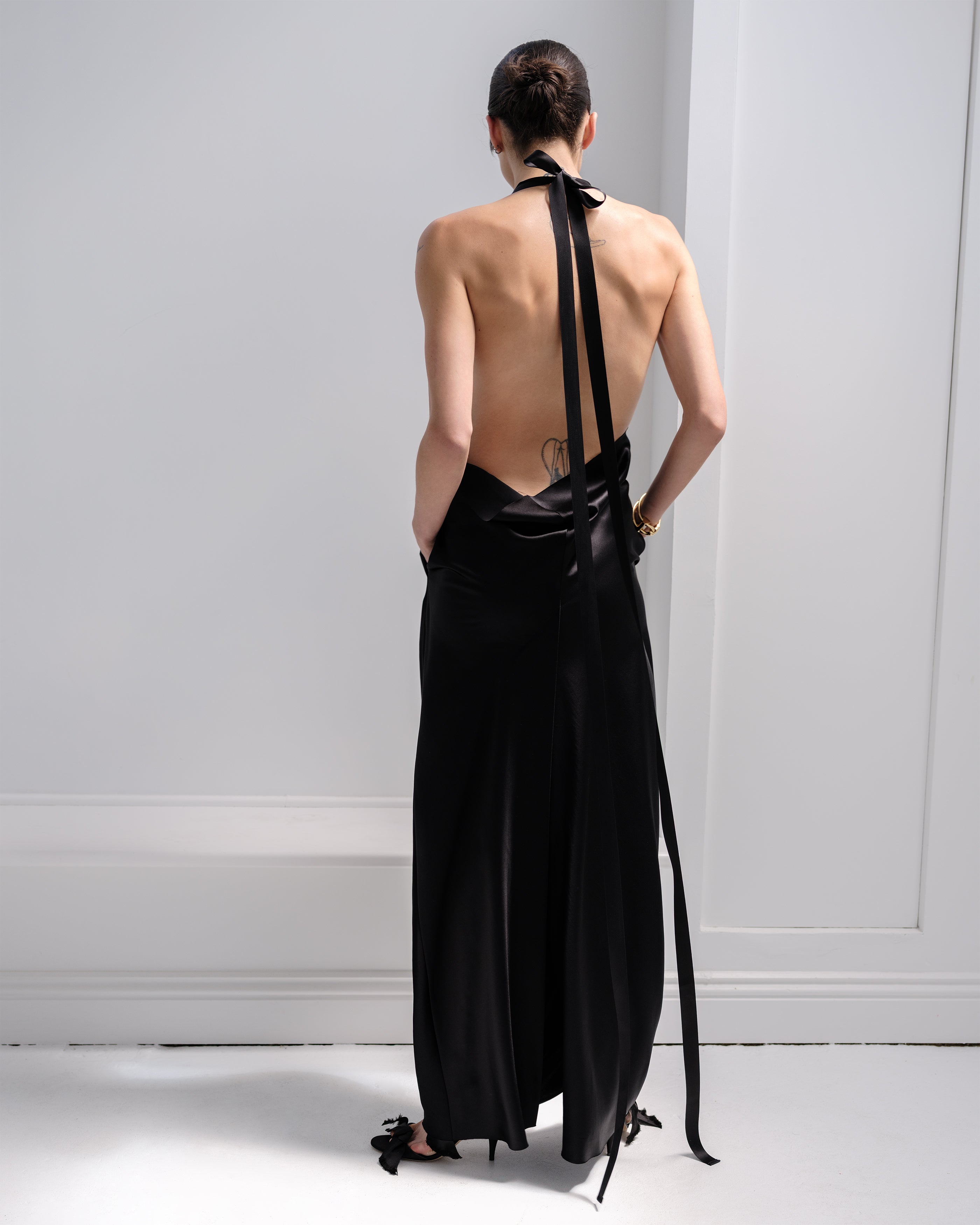 Woman wearing a black Common Hours backless halter neck gown with long ribbon straps against a white wall.