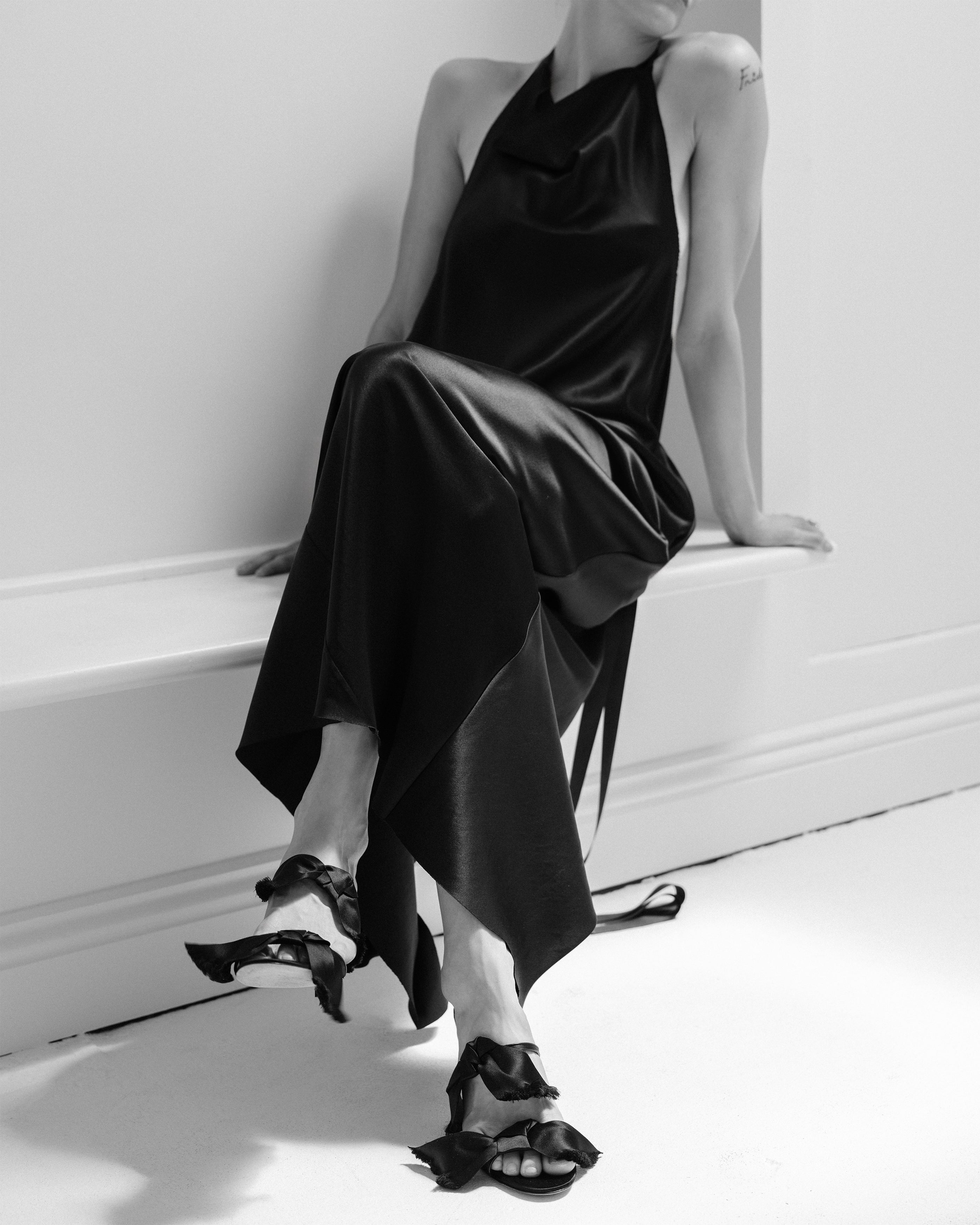 Woman in a black satin halter neck Common Hours dress sitting on a white bench.