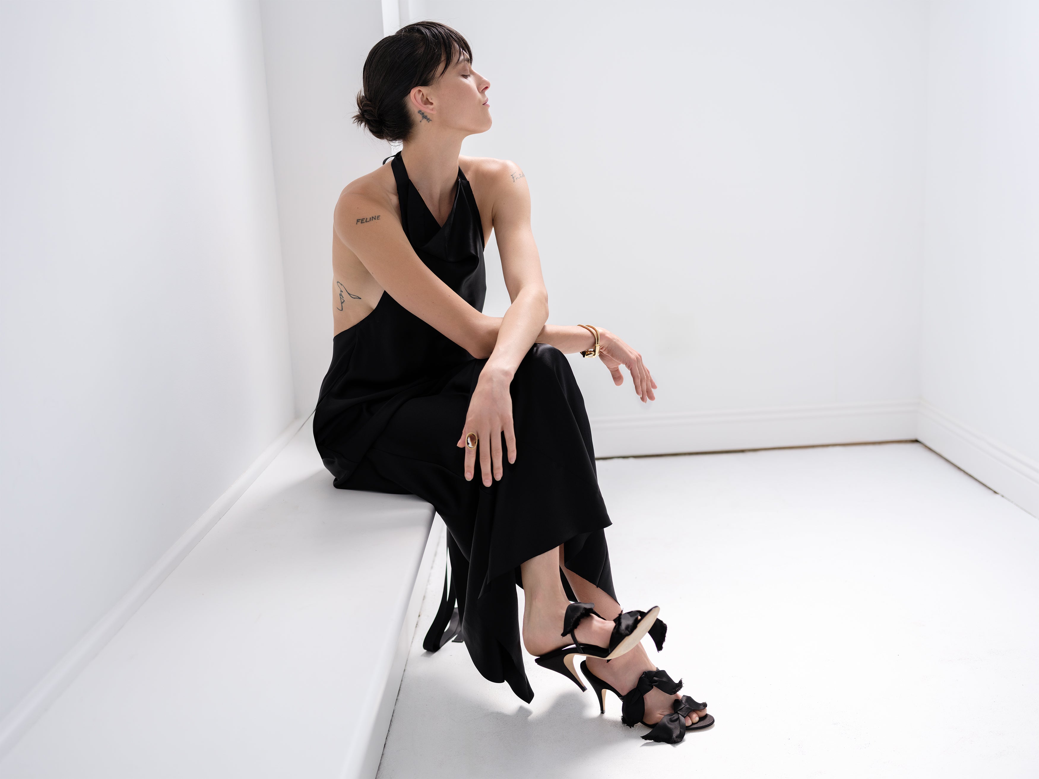 Woman in a black Common Hours silk halter dress and high ribbon heels sitting on a white bench in an empty room with a white background.