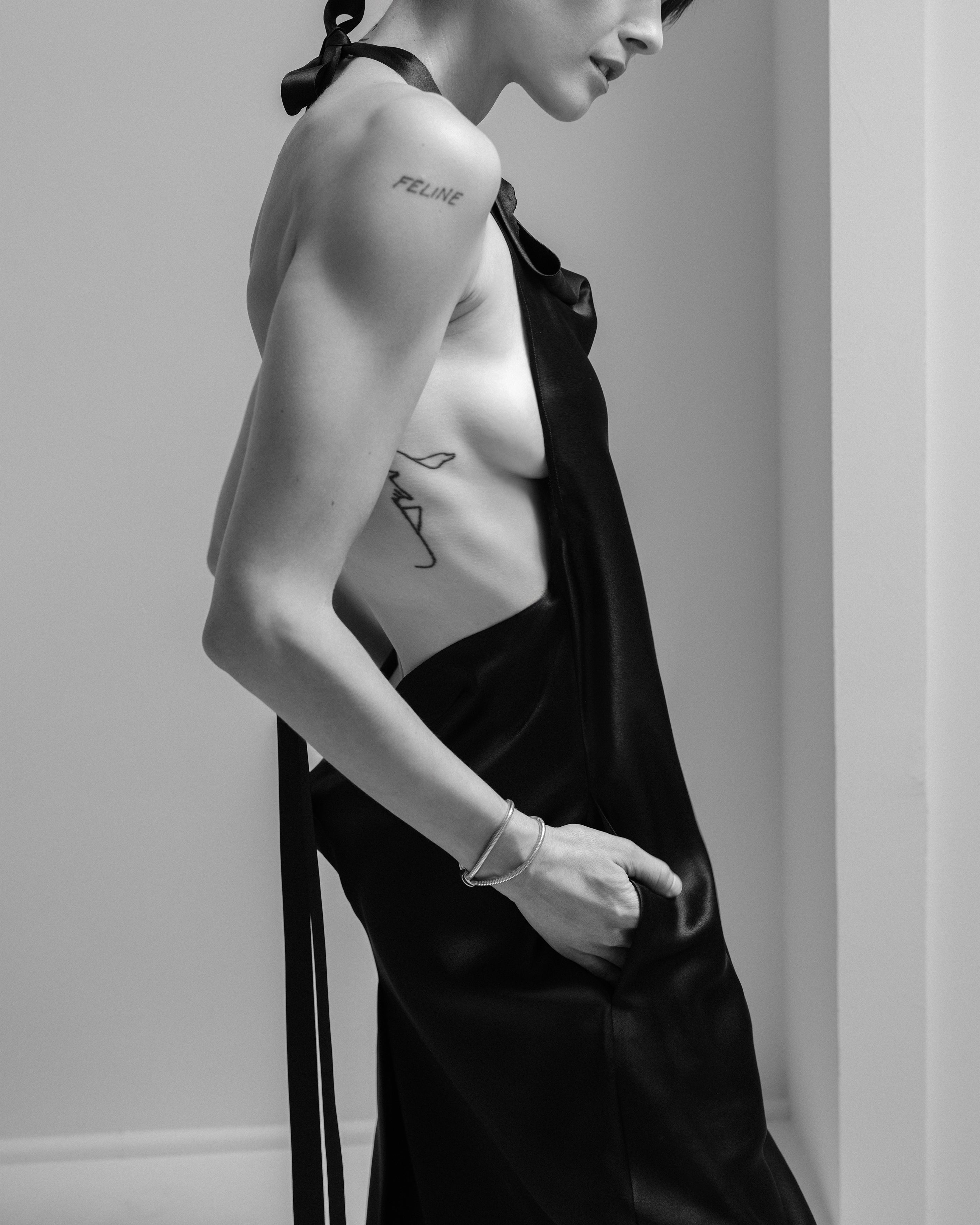 Woman wearing a black Common Hours halter neck silk dress with long ribbon straps and visible tattoos on a plain background