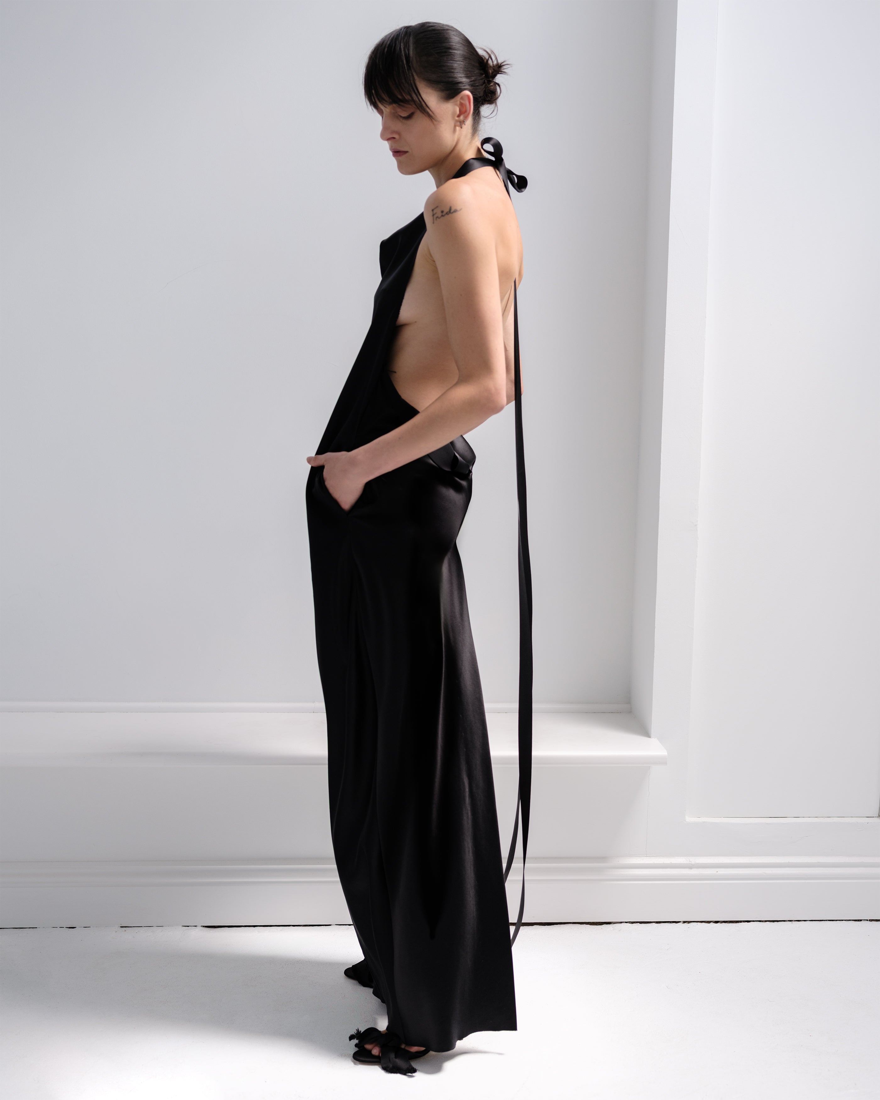 Woman wearing a black Common Hours backless dress with a long ribbon straps against a white wall.