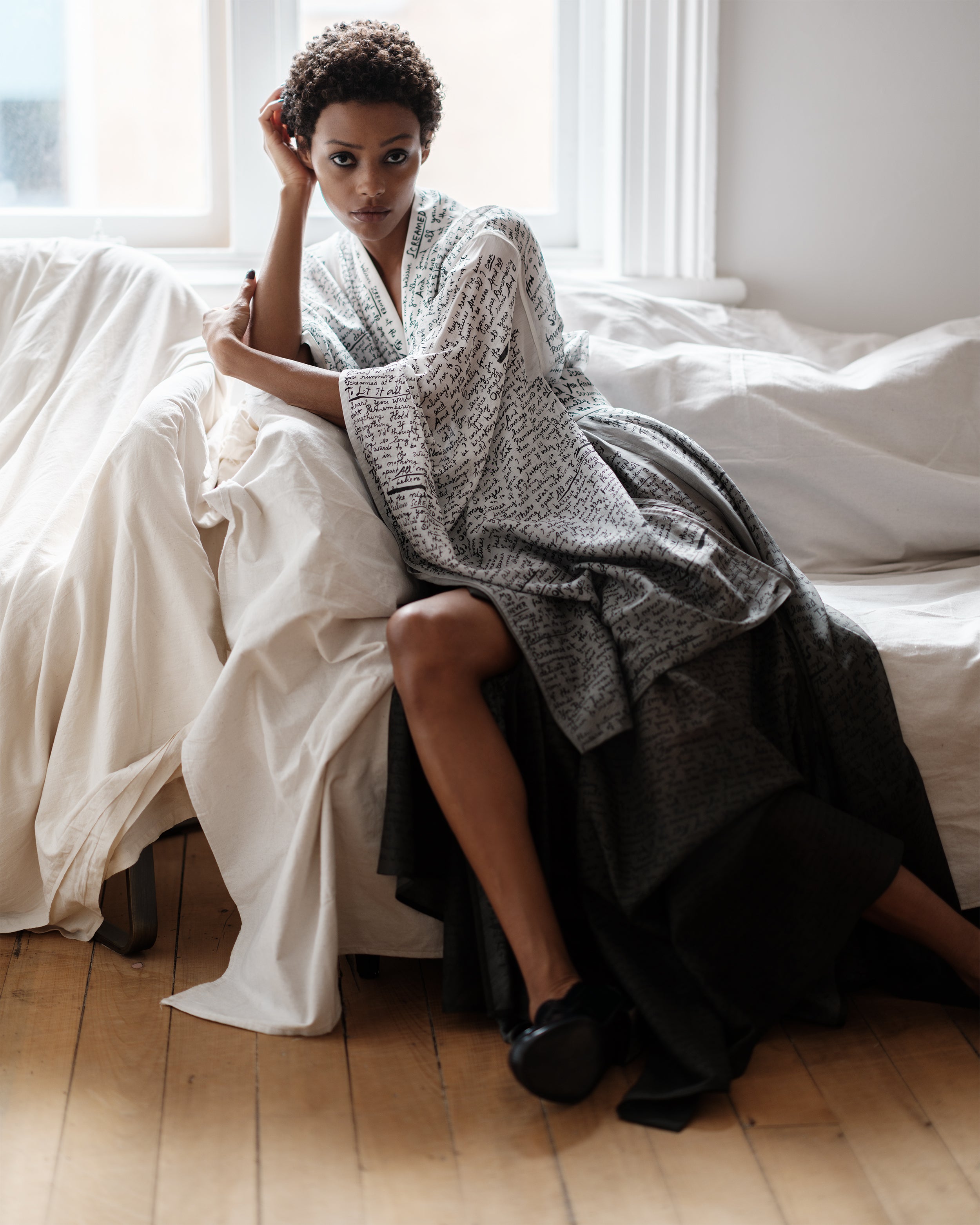 Woman sitting on a bed wearing a gradient Common Hours wrap-around robe dress with printed and hand-embroidered lyrics in a bright room.