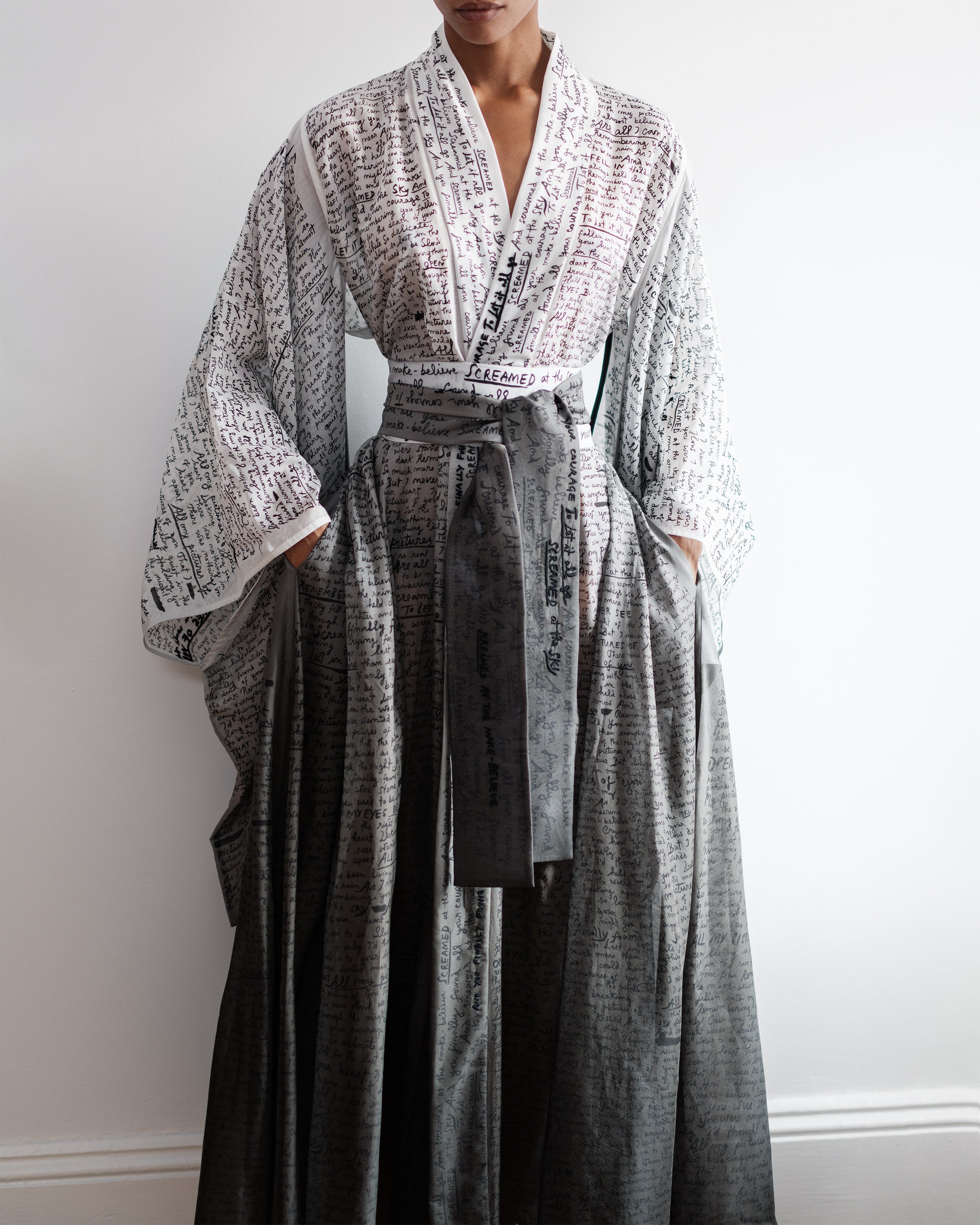 Woman wearing a long Common Hours robe dress with printed and hand-embroidered lyrics against a plain background