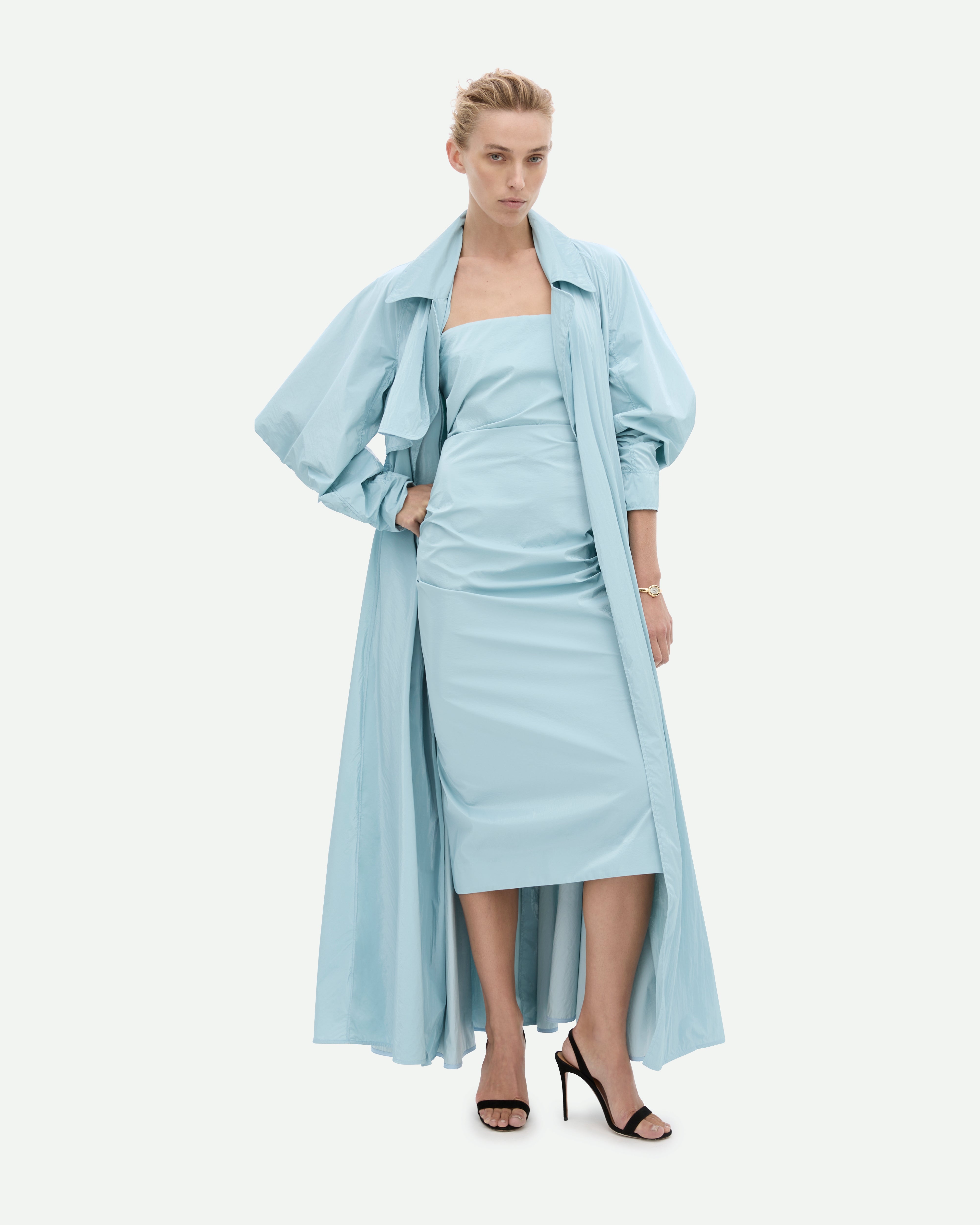 A woman wearing a strapless, sky blue, mid-length dress with a fitted silhouette, Tabloid dress by Common Hours. Coat overlaid