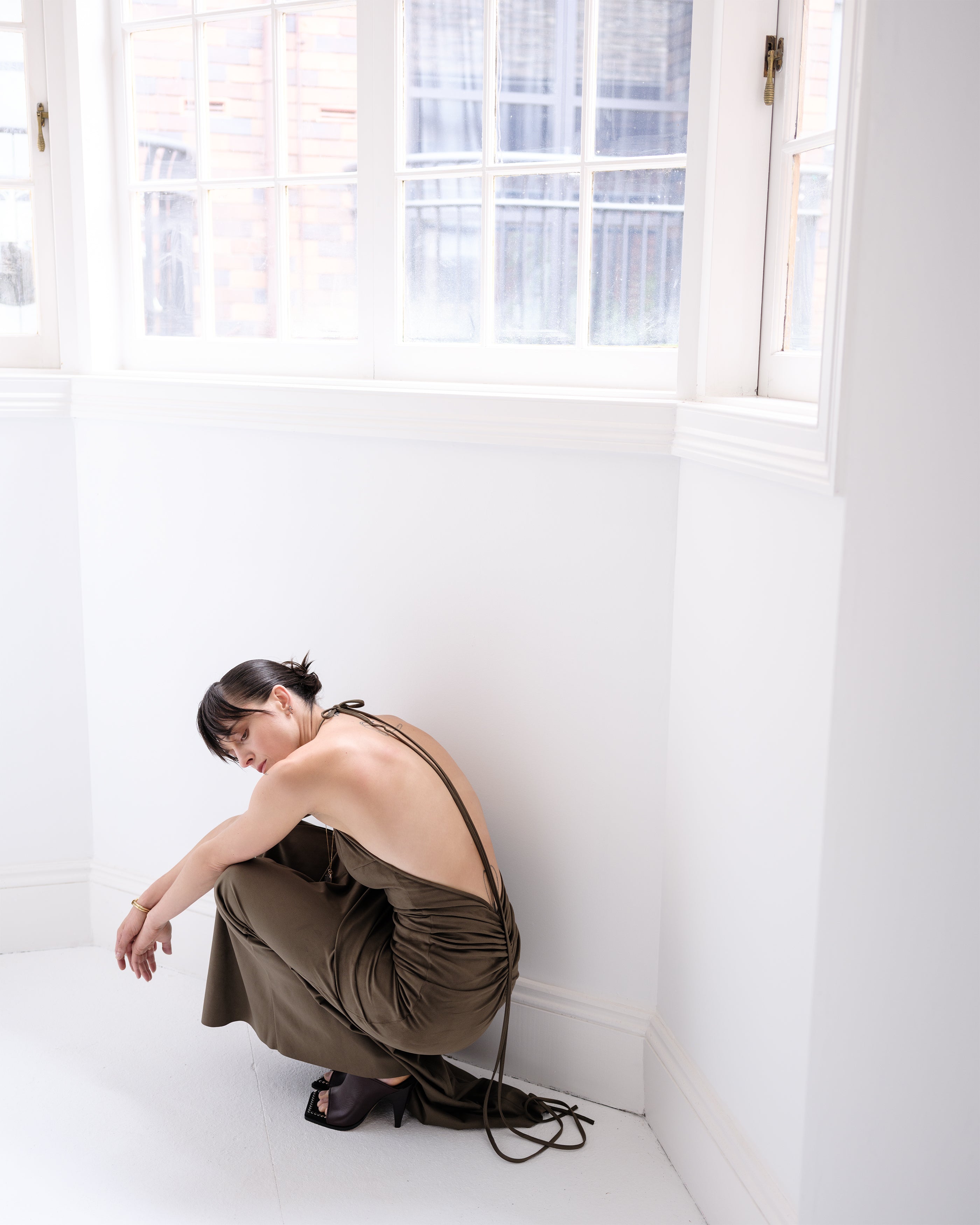 Woman sitting on white floor with a khaki open back Bluet dress from Common Hours.