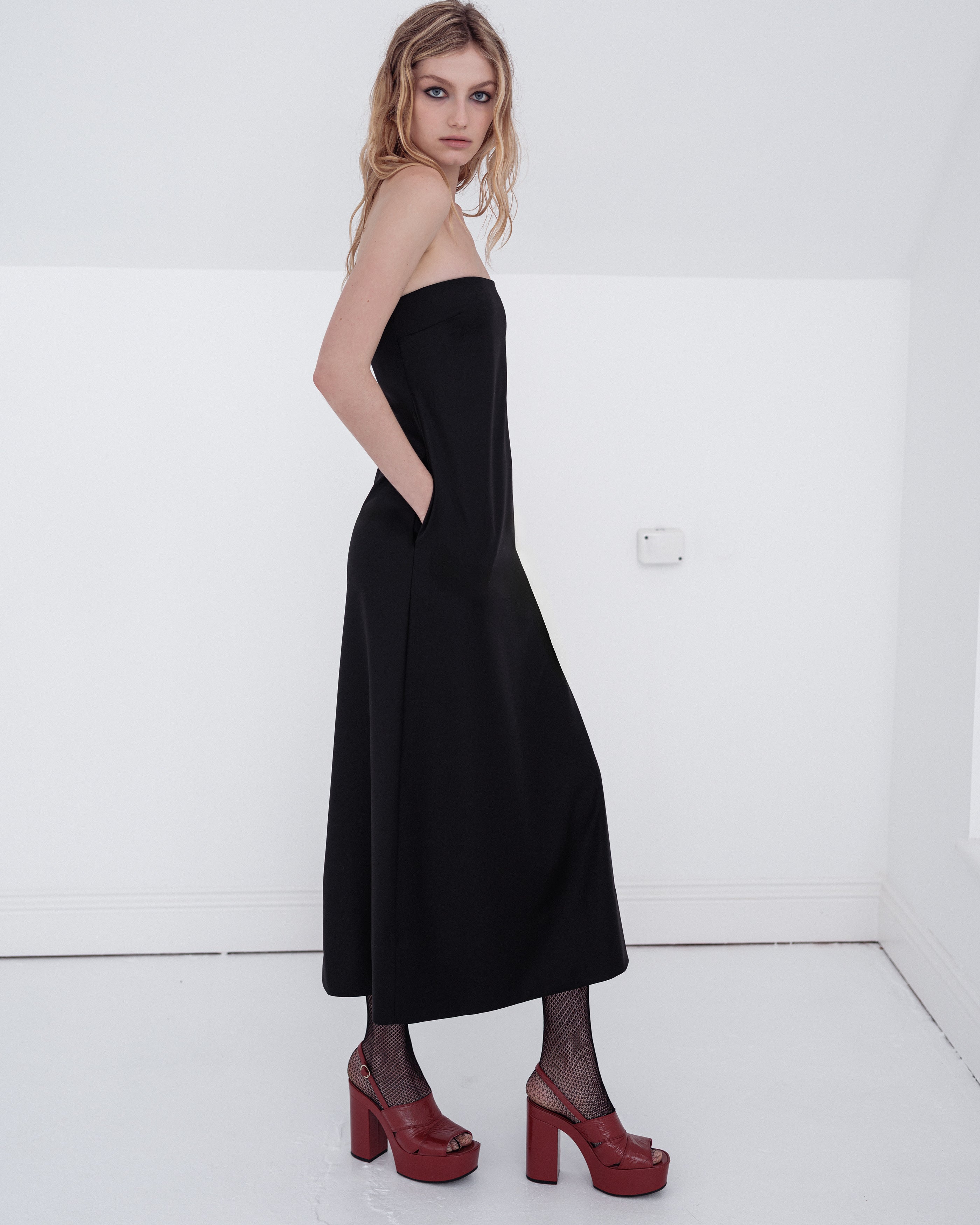 Woman wearing a classic black Common Hours strapless dress with red high-heeled shoes on a white background