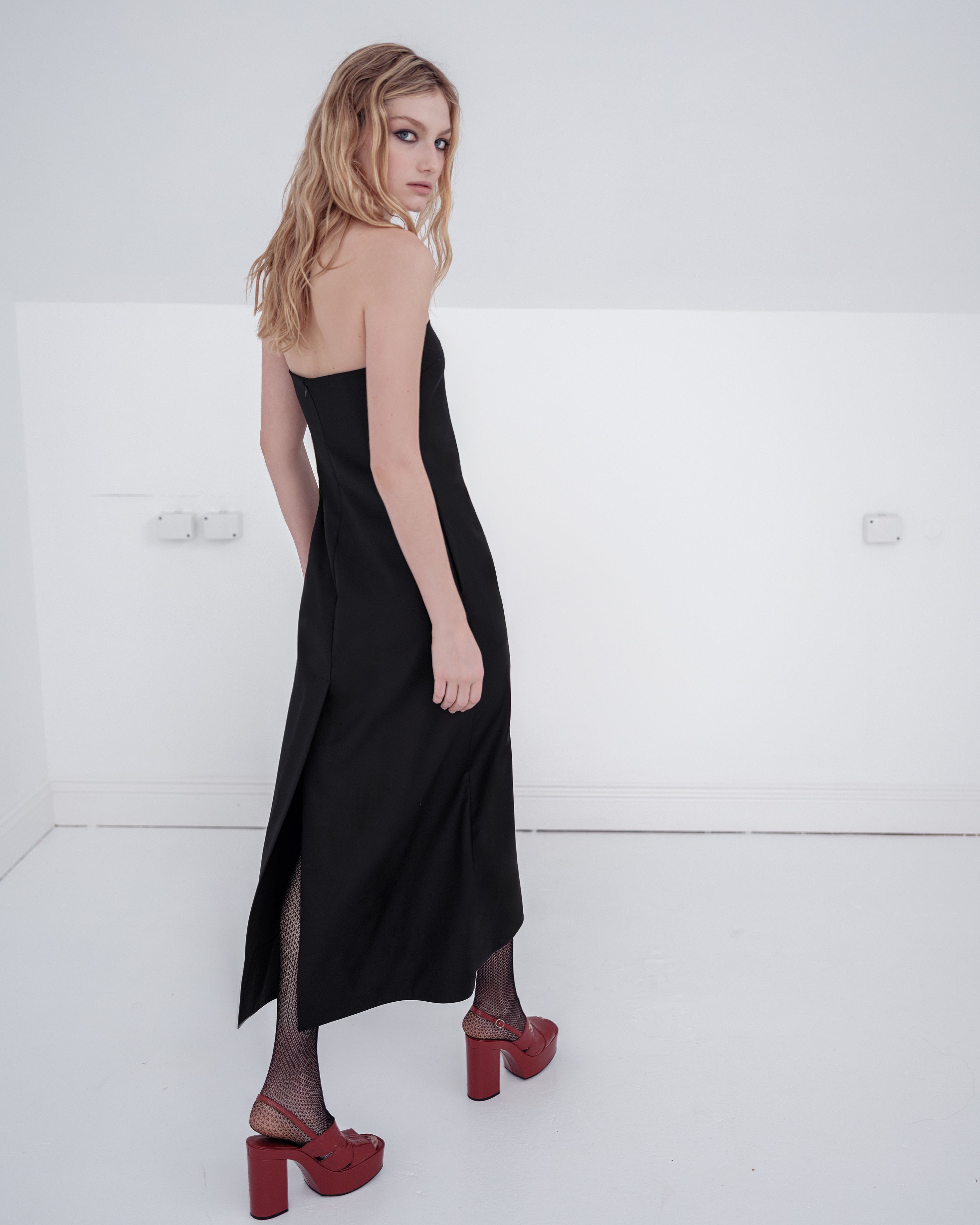 Woman wearing a black strapless Common Hours dress with red shoes on a white background