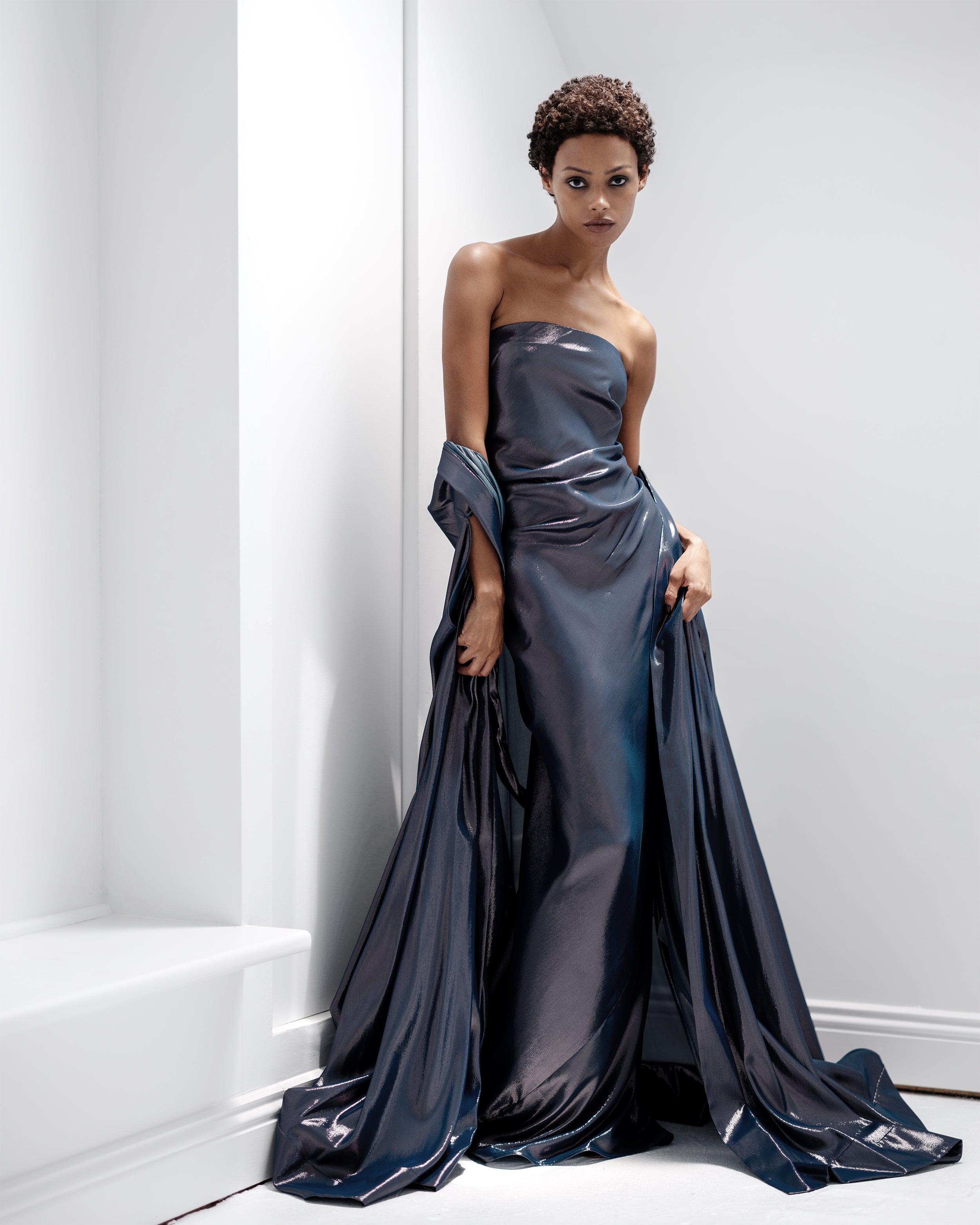 Woman in a strapless, dark blue Common Hours evening gown against a white wall.