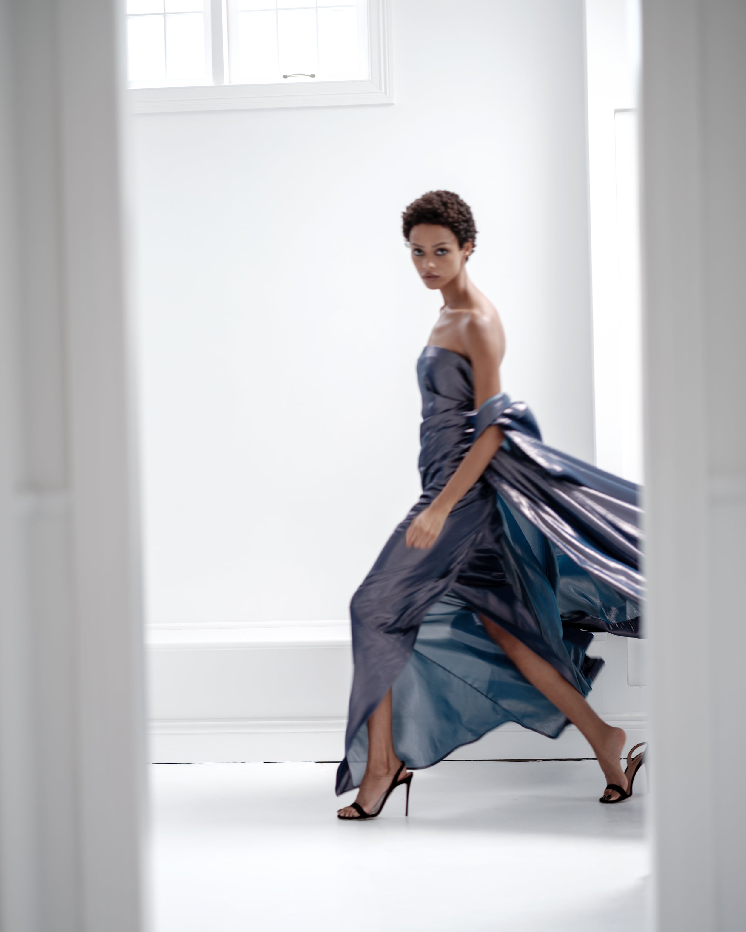Woman in a strapless blue Common Hours gown standing in a bright, minimalistic room.