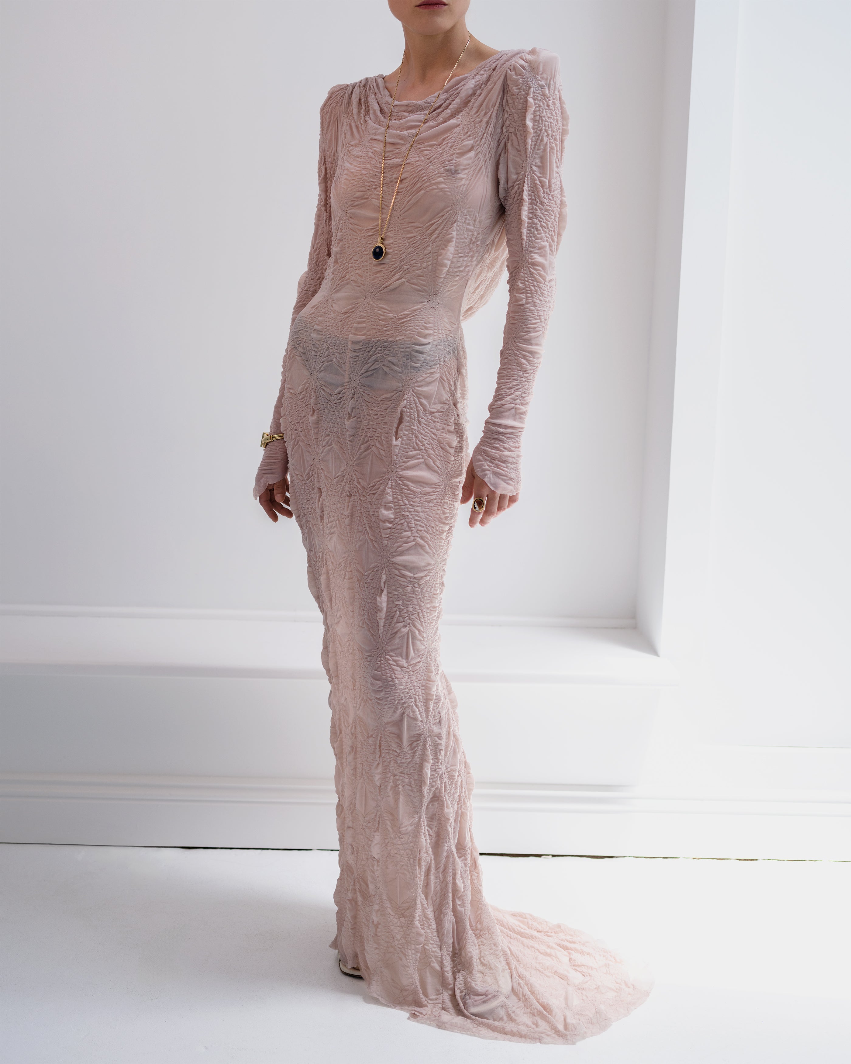 Woman wearing a long, see-through blush Intention Common Hours dress with a white background.