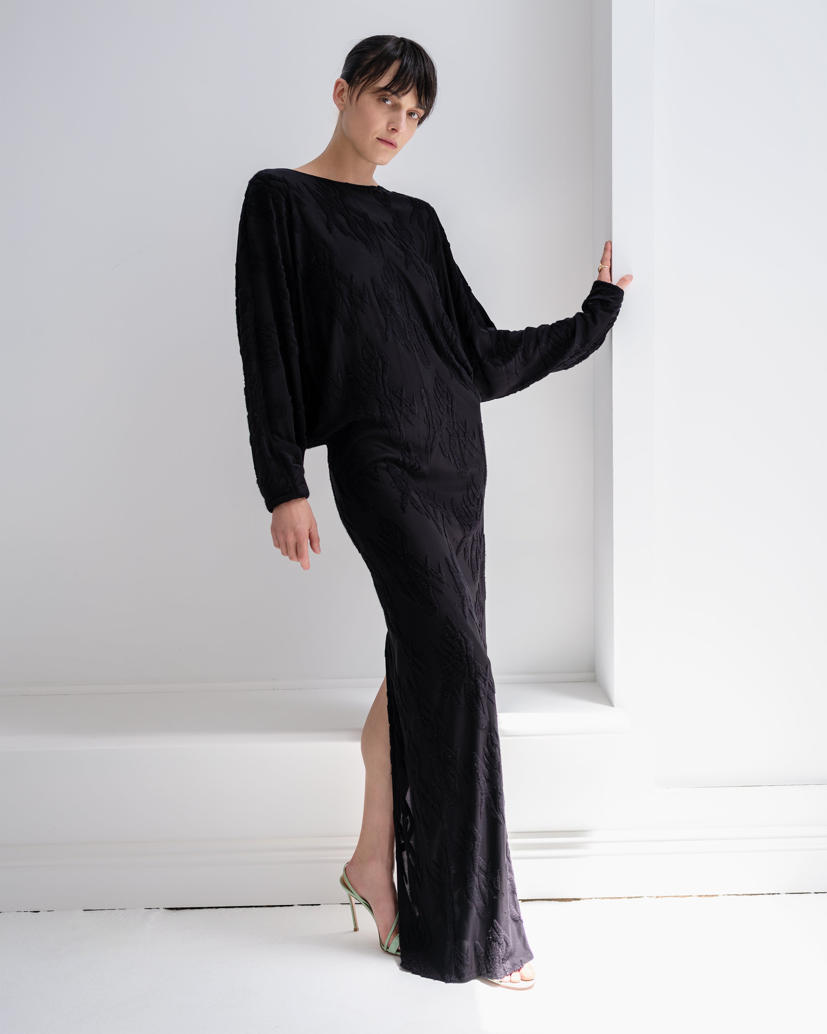 Woman wearing a black Common Hours Offset dress with a slit on a white background.