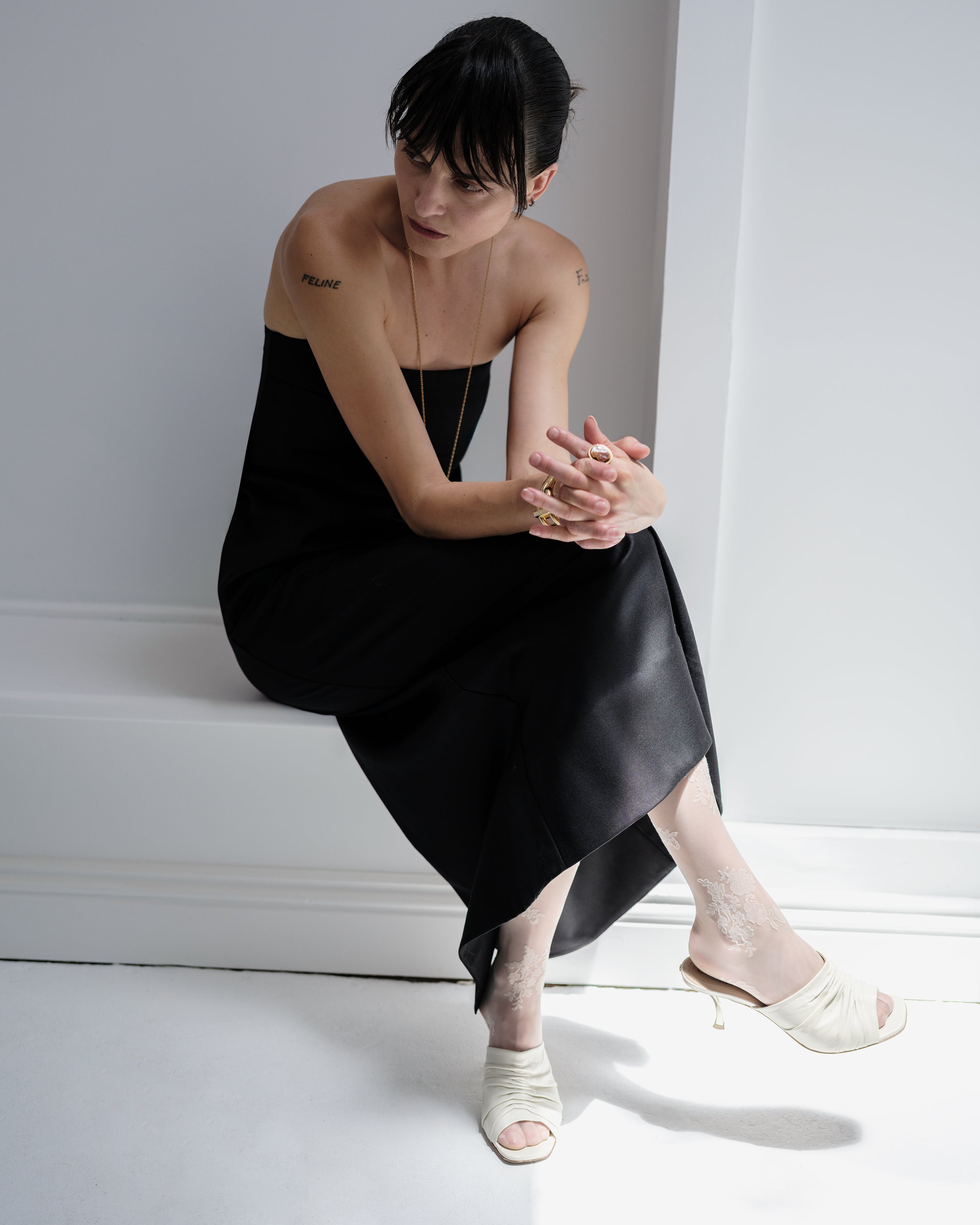 Woman in a black Common Hours strapless dress sitting on a white floor with a neutral background.