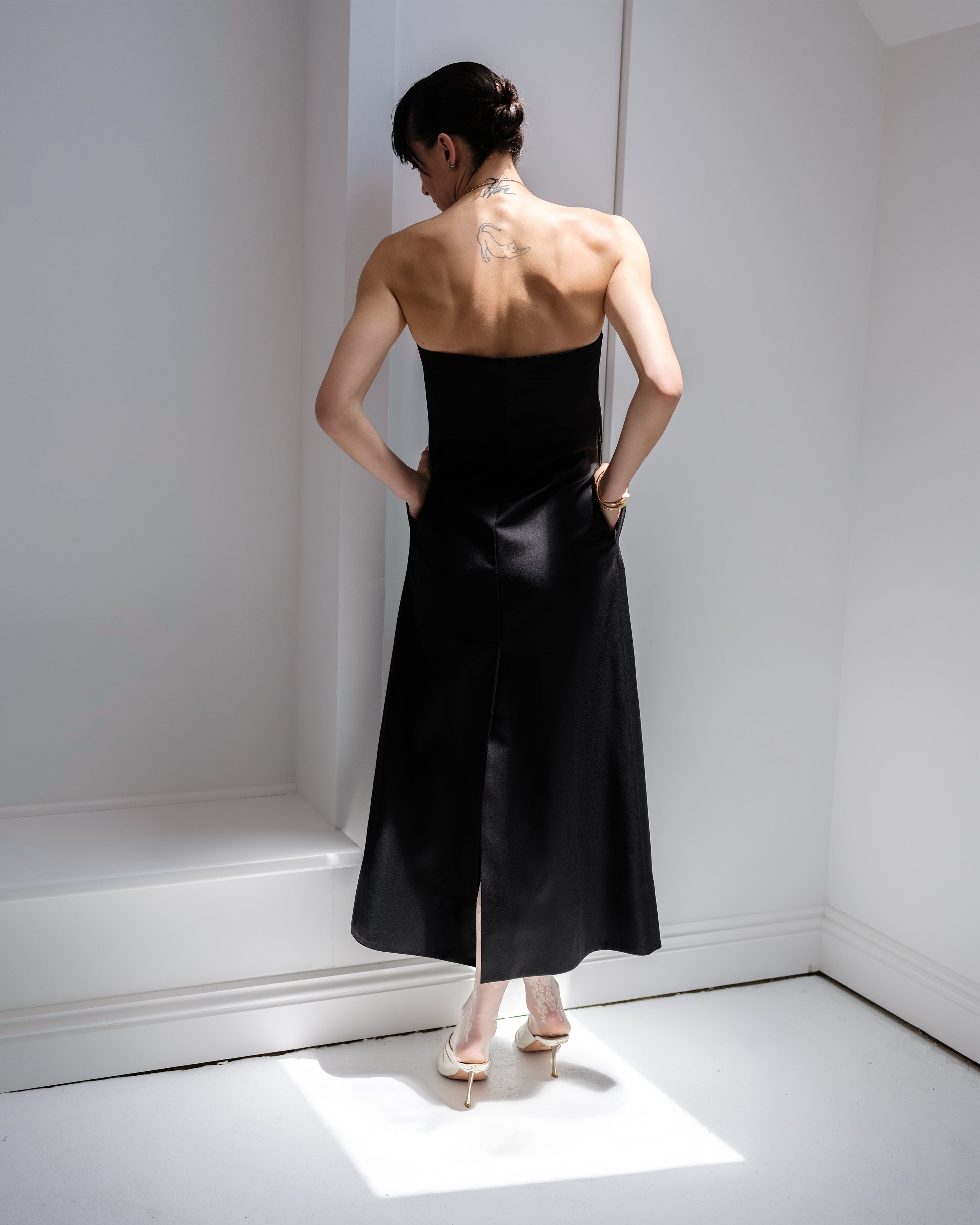 Woman wearing a classic black Common Hours strapless dress standing in a minimal white room.