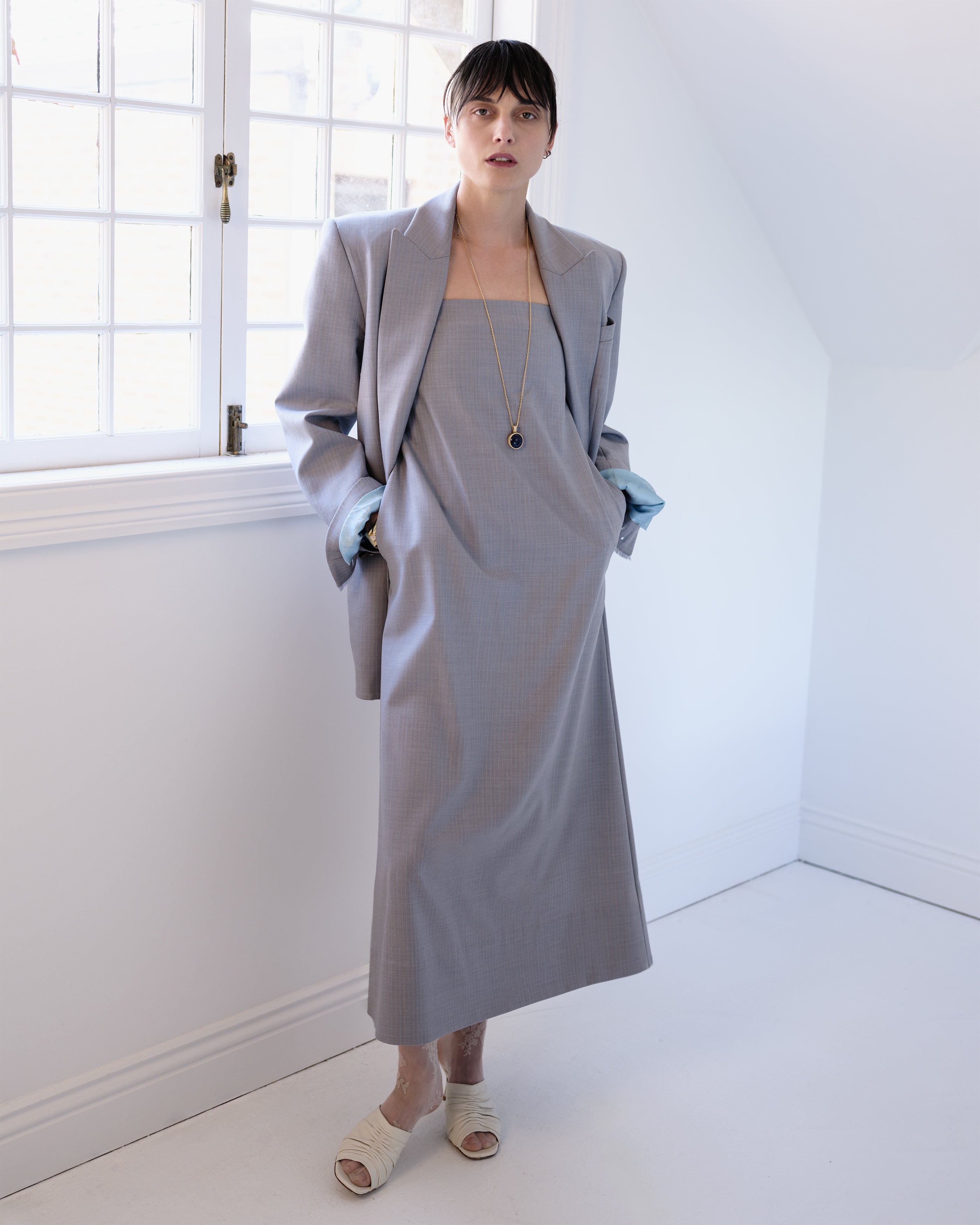 Woman wearing a gray Common Hours strapless dress and matching blazer in a minimal white room.