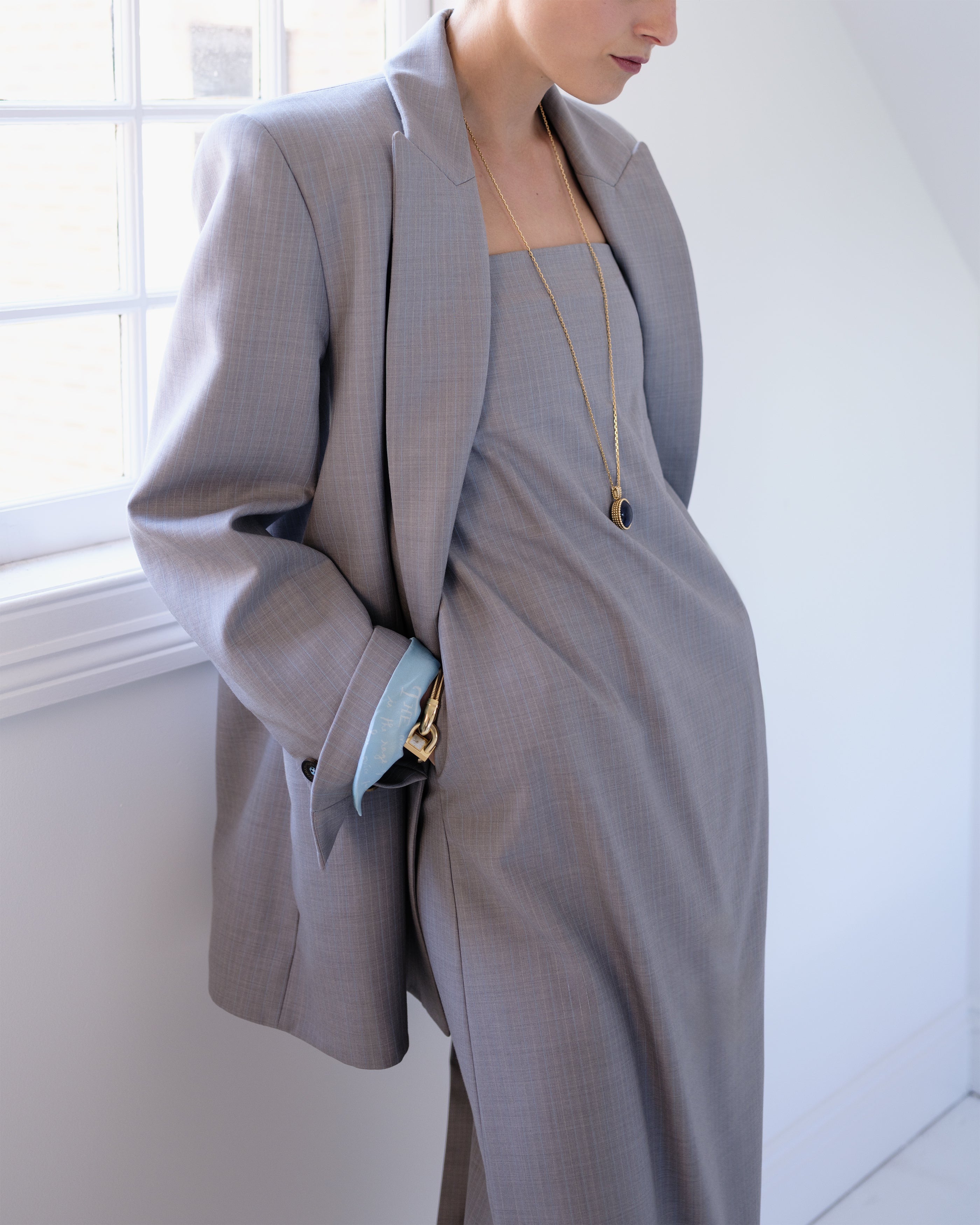 Woman wearing a gray Common Hours strapless suiting dress with matching blazer standing against a window.