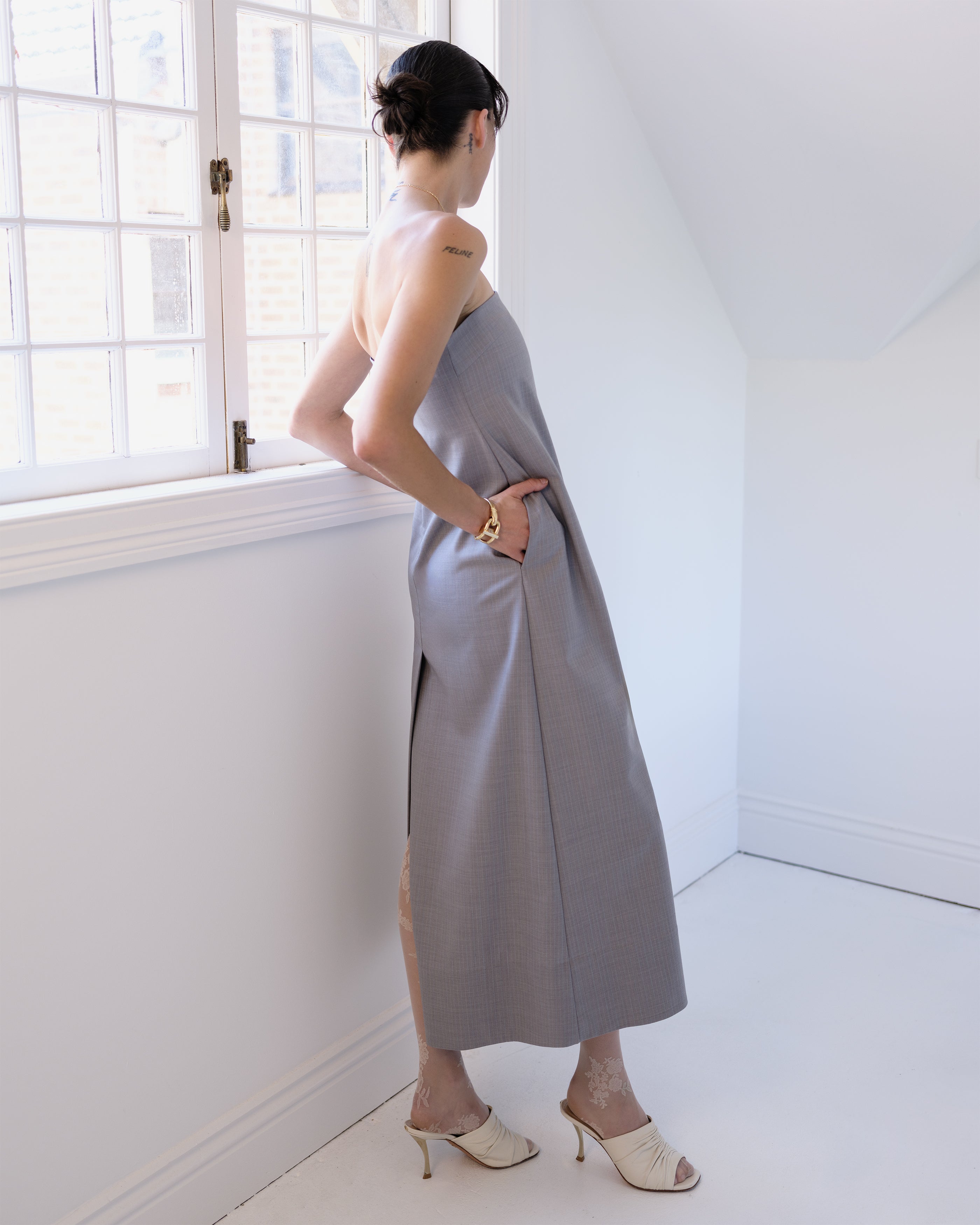 Woman in a gray Common Hours strapless dress standing in a bright room with white walls and a window.