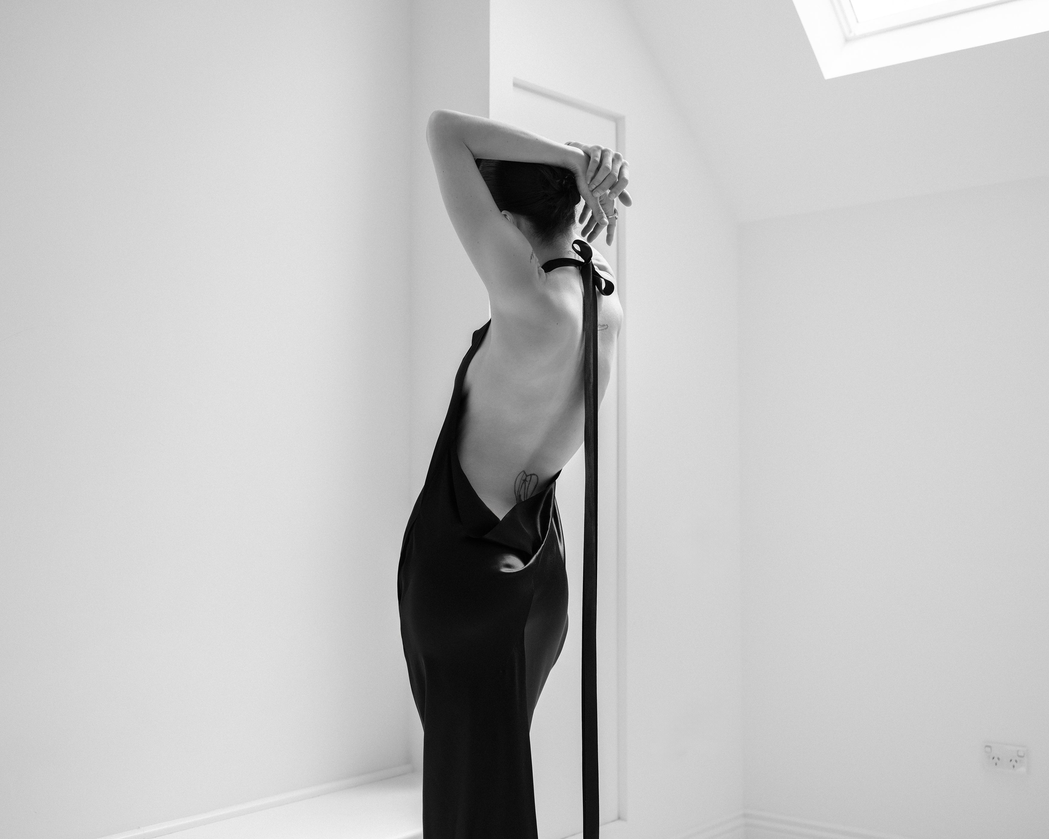 Woman in a black Common Hours halter neck black dress stretching in a minimalistic room.