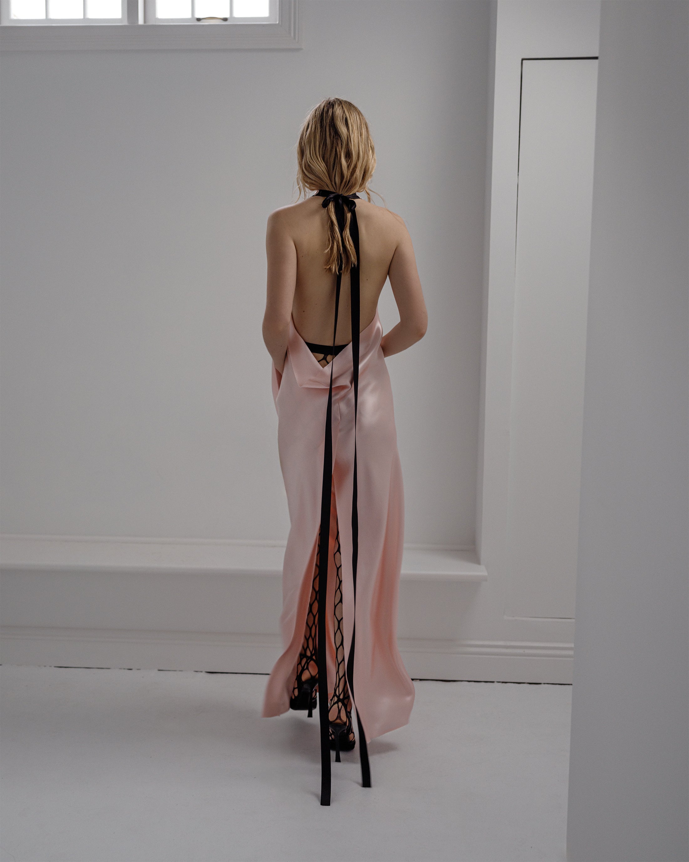 Woman in a pink Common Hours backless satin gown with black ribbon details standing in a minimalistic room.