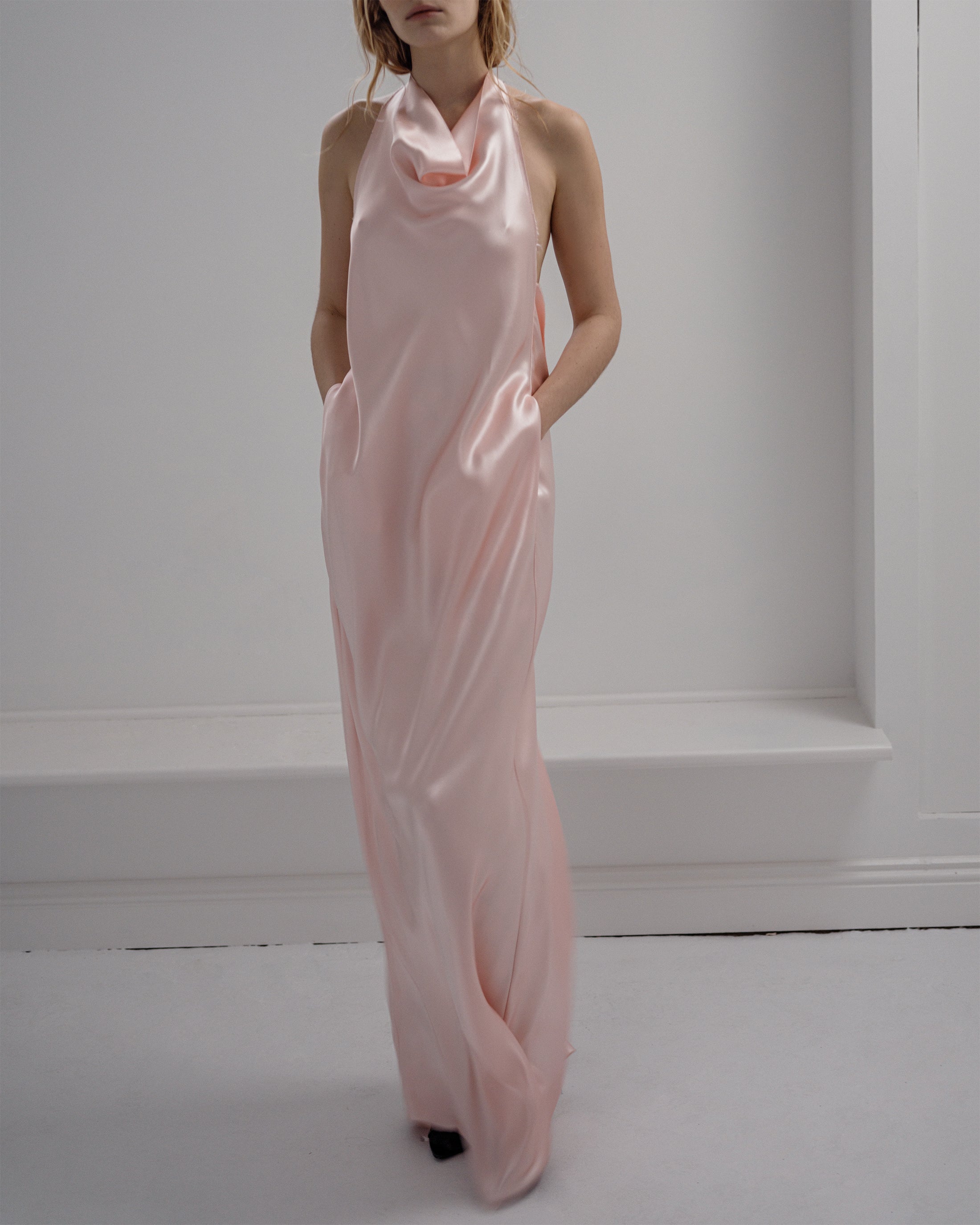 Woman wearing a long Common Hours pink satin draped dress against a white wall.