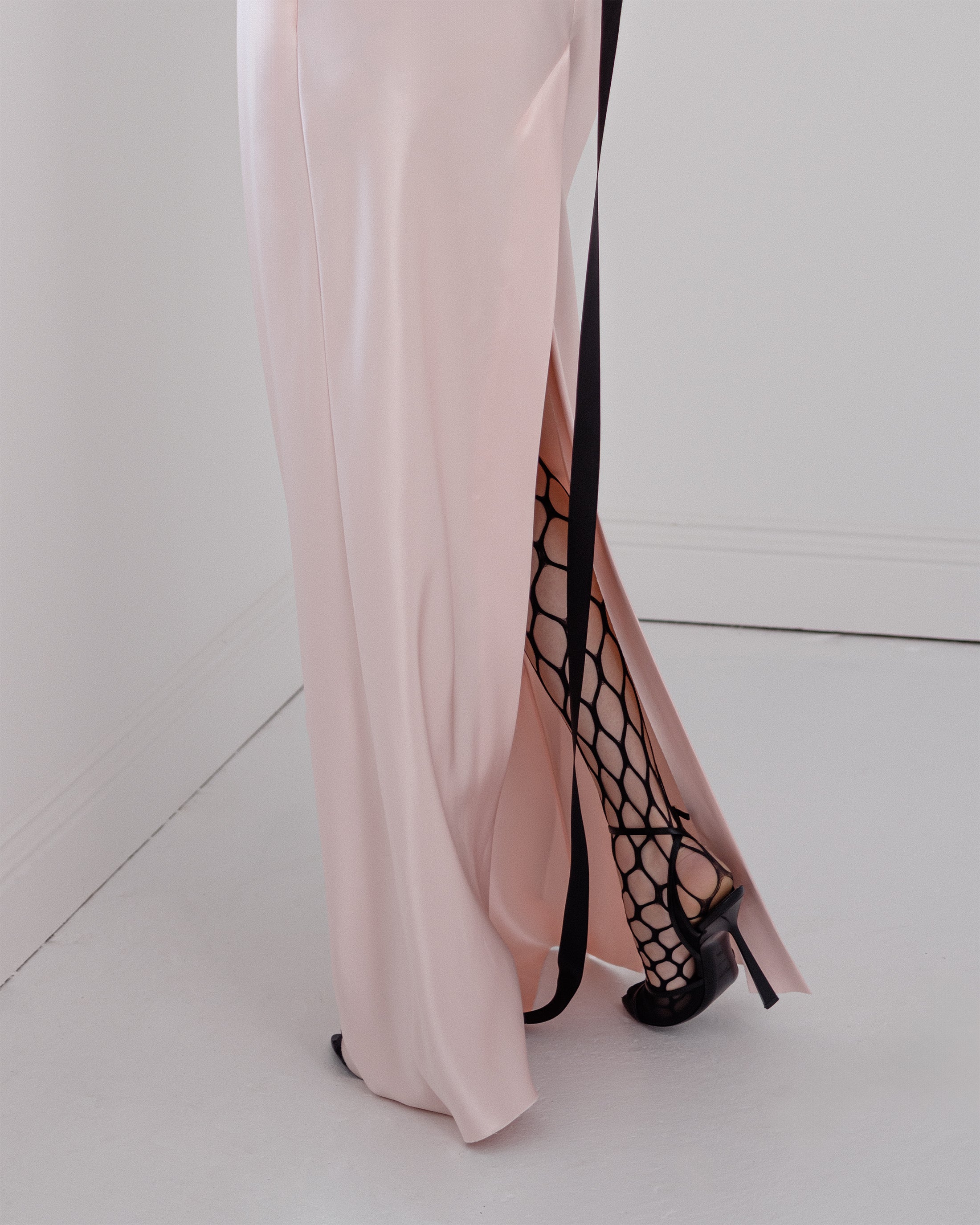 Woman wearing a pink Common Hours satin dress with a high slit and black strappy heels on a white floor.