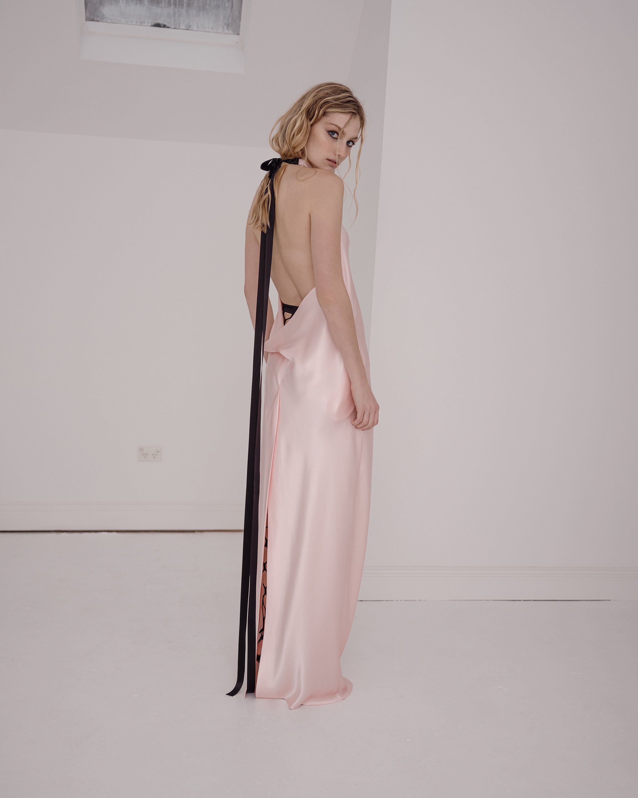 Woman wearing a backless pink silk Common Hours evening gown with black ribbons against a white background