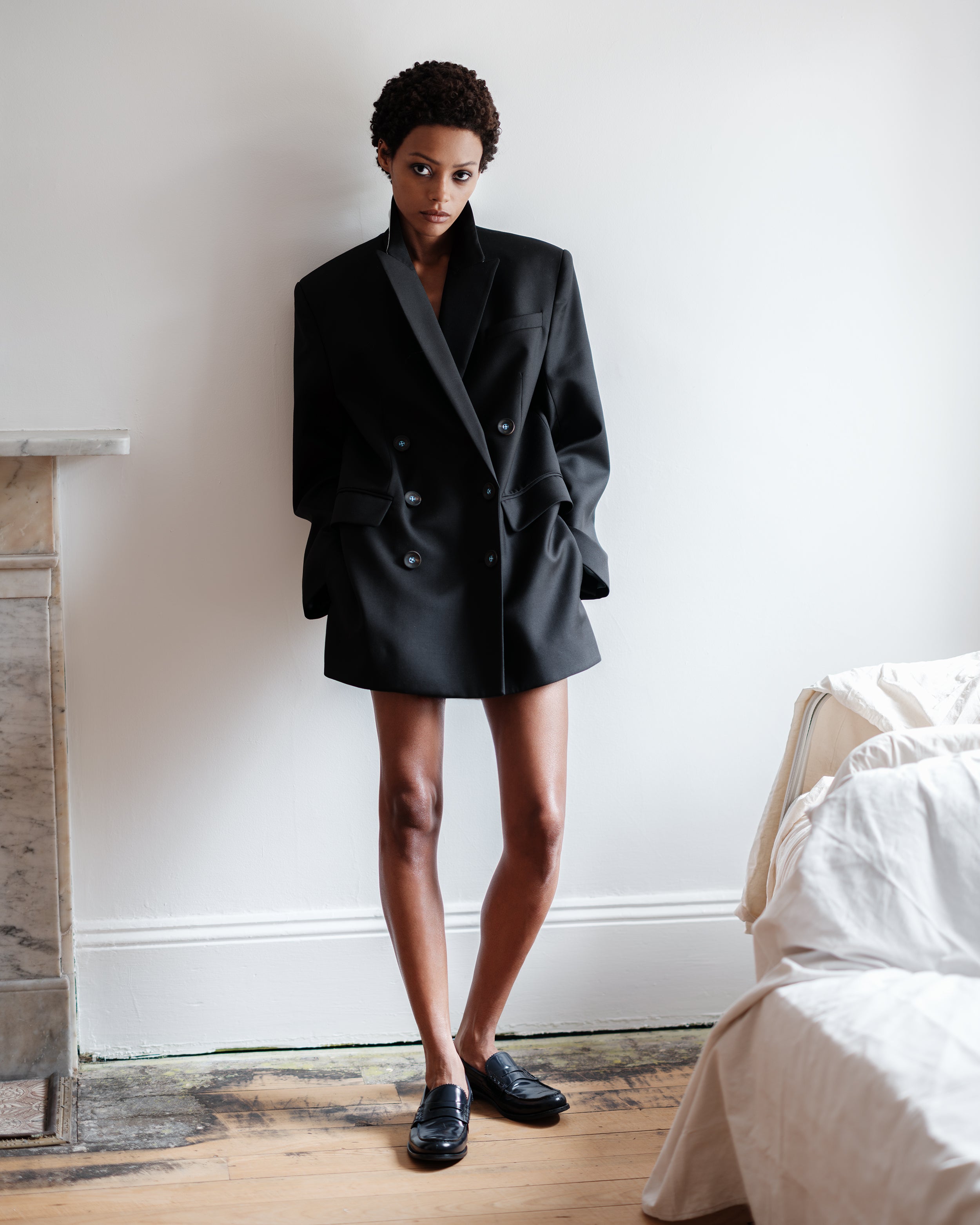 Woman wearing a black oversized Common Hours blazer dress in Italian wool, standing in a room with a bed and fireplace.