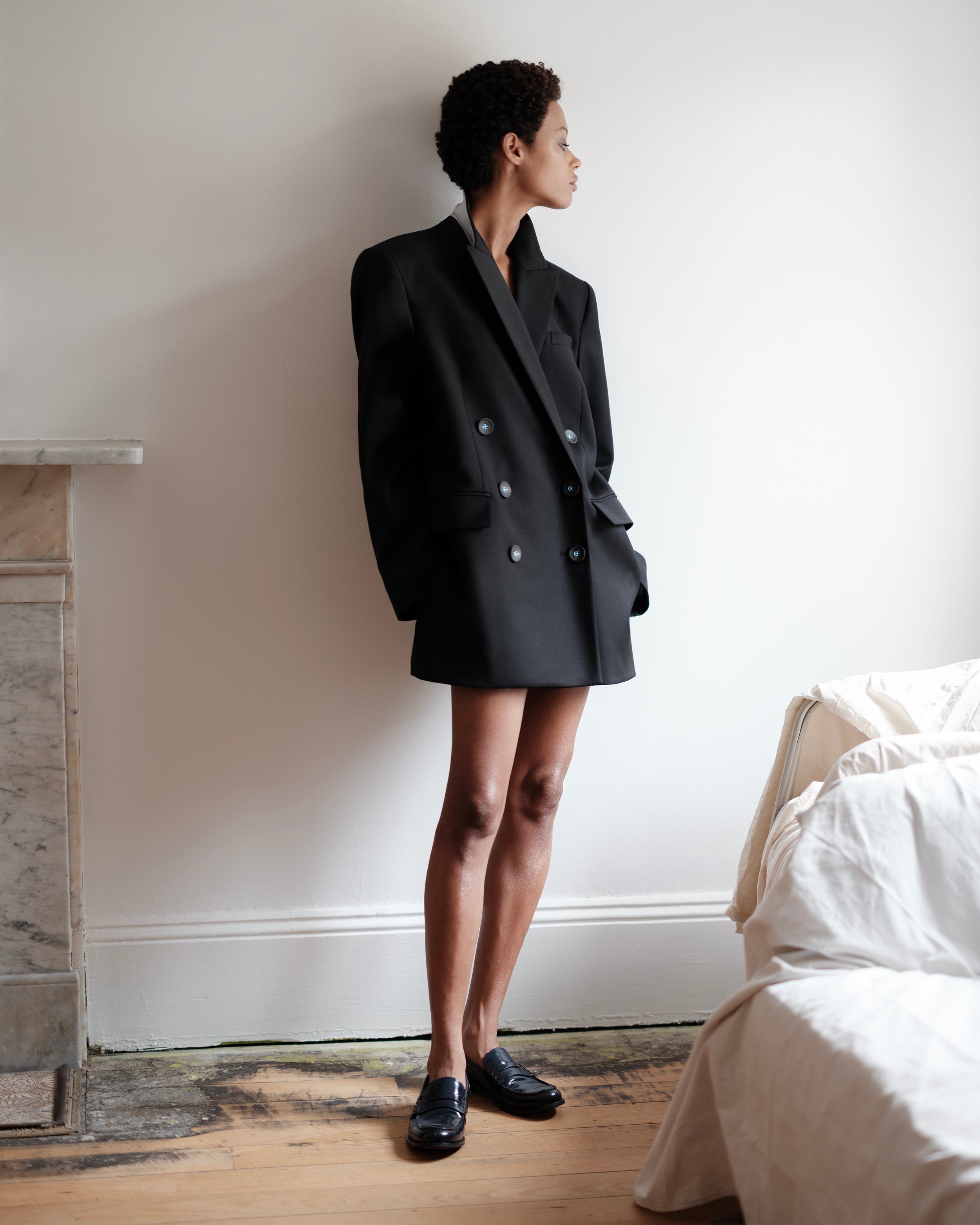 Woman wearing a black oversized Common Hours blazer dress standing in a room with a bed and fireplace.