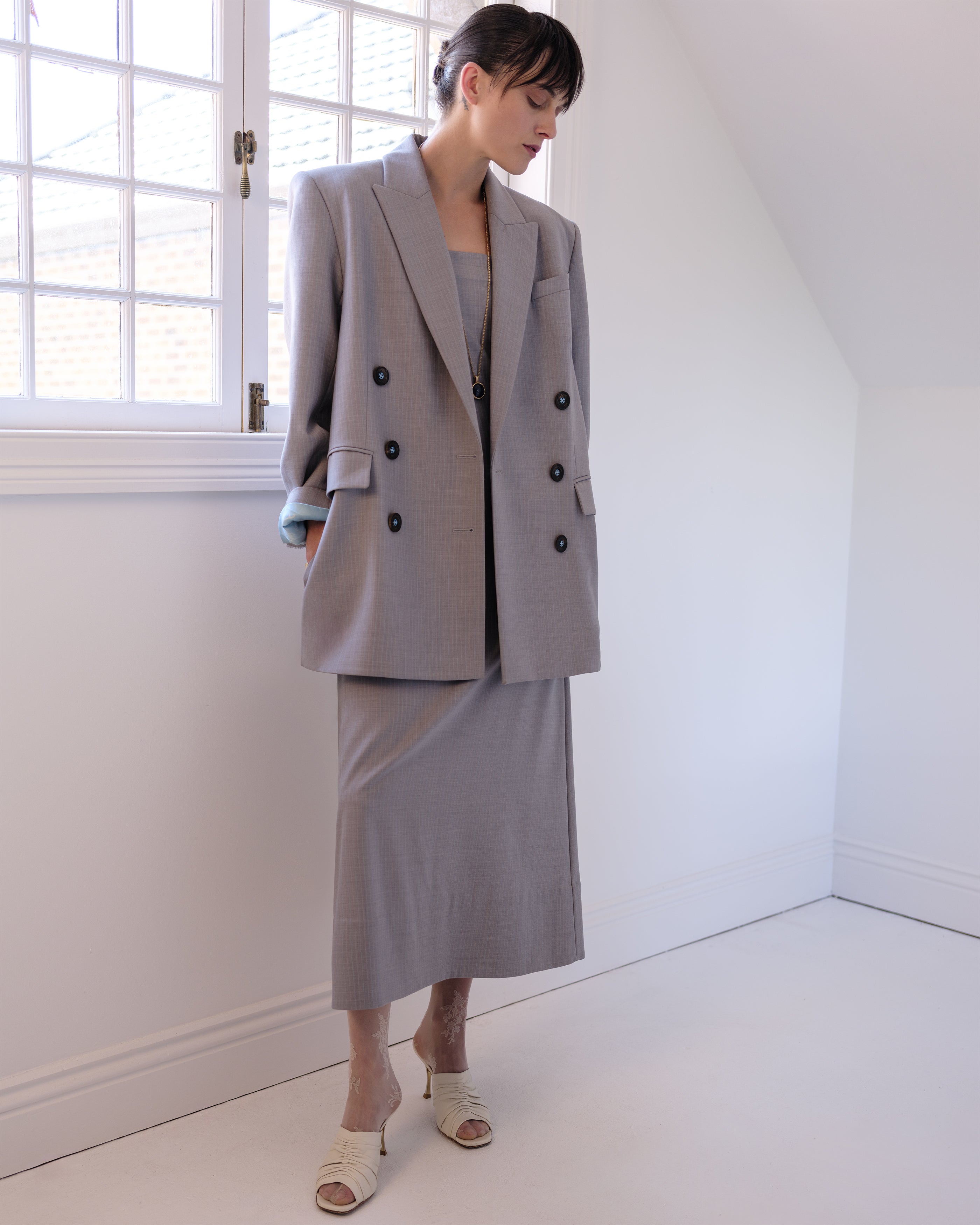 Woman wearing a gray Common Hours blazer and matching strapless dress standing in a bright room with white walls and floor.