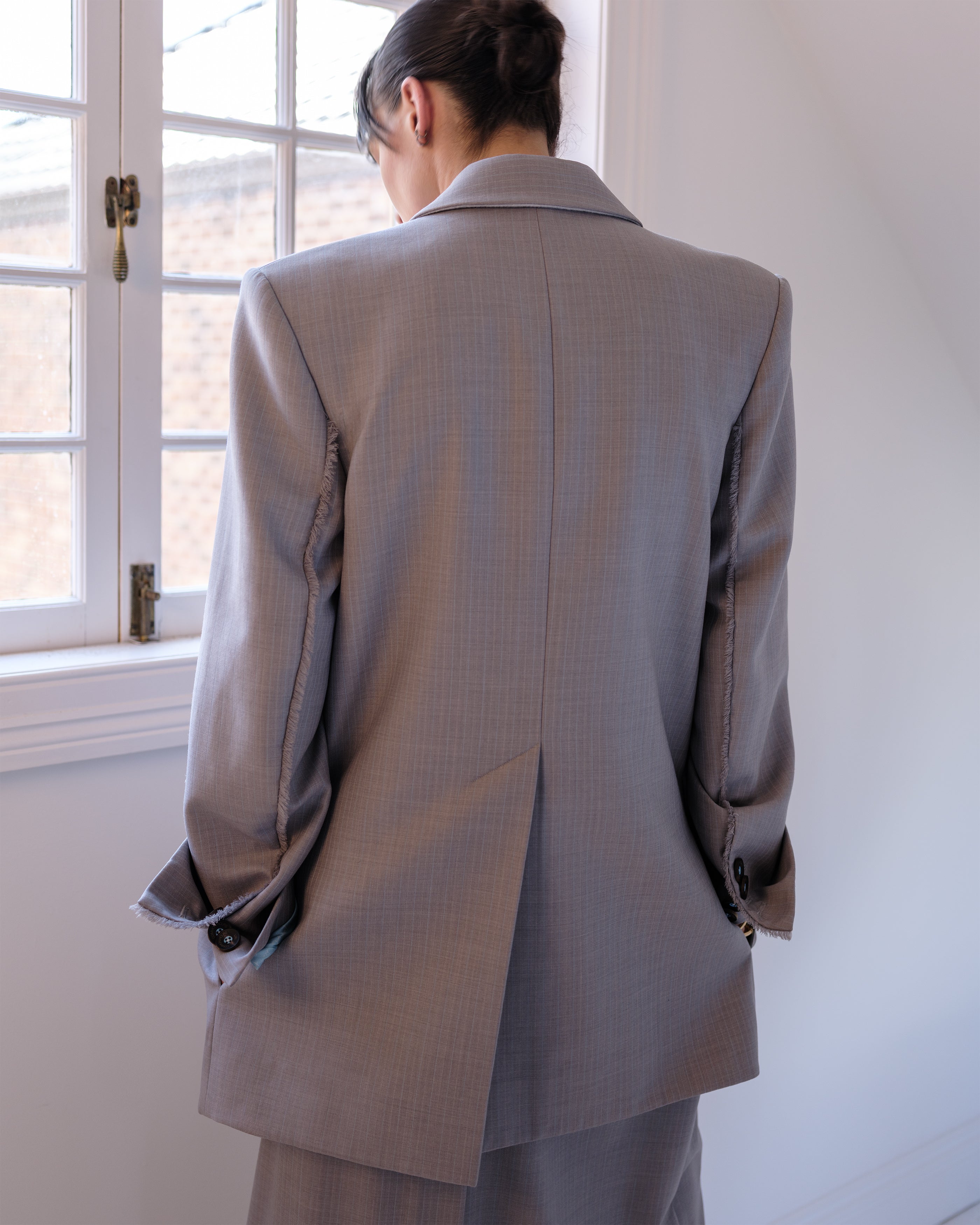 Woman wearing a Common Hours gray oversized suiting jacket dress with sharp shoulders standing in front of a window.