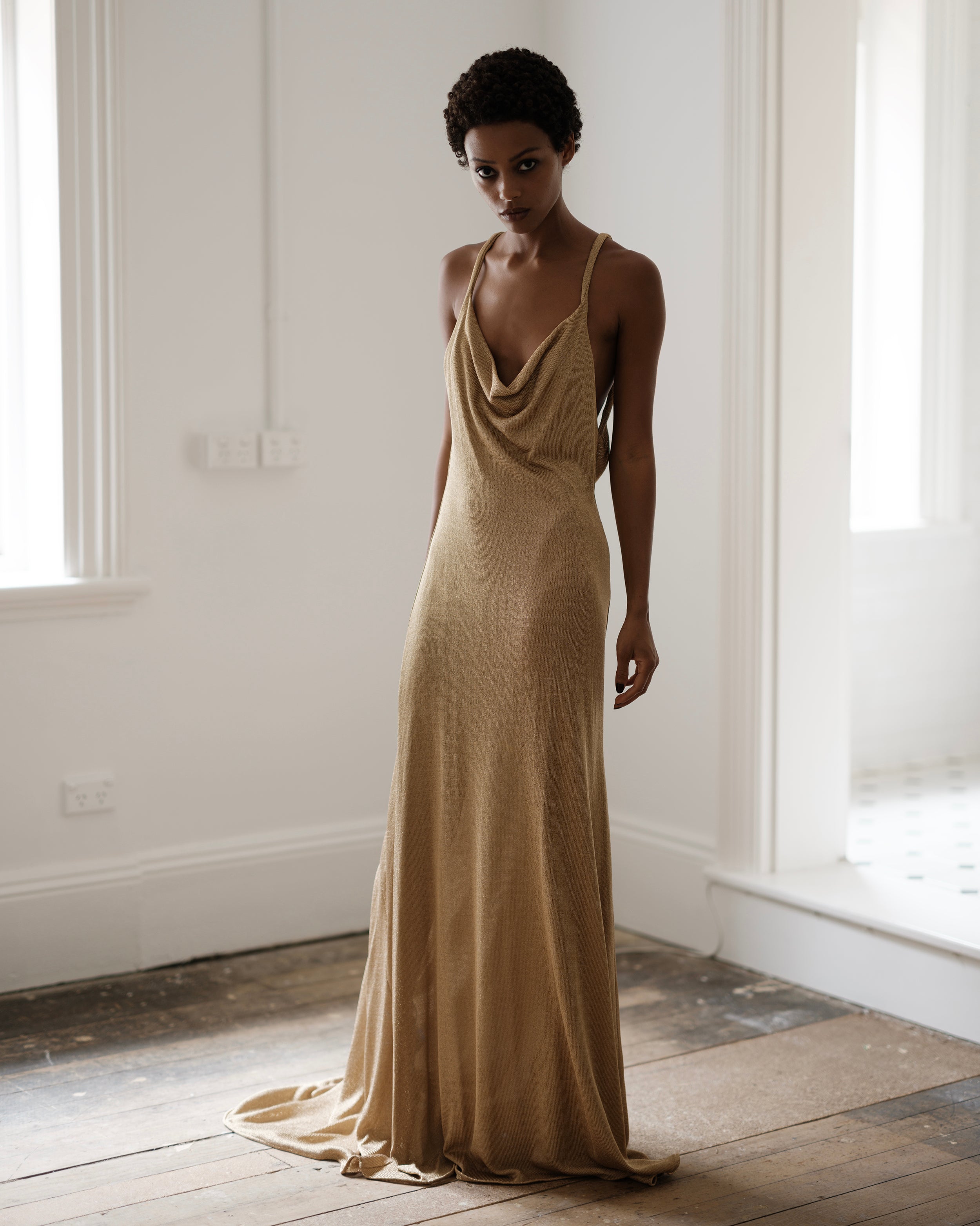 Woman wearing a metallic gold Common Hours evening gown in a bright room with large windows.