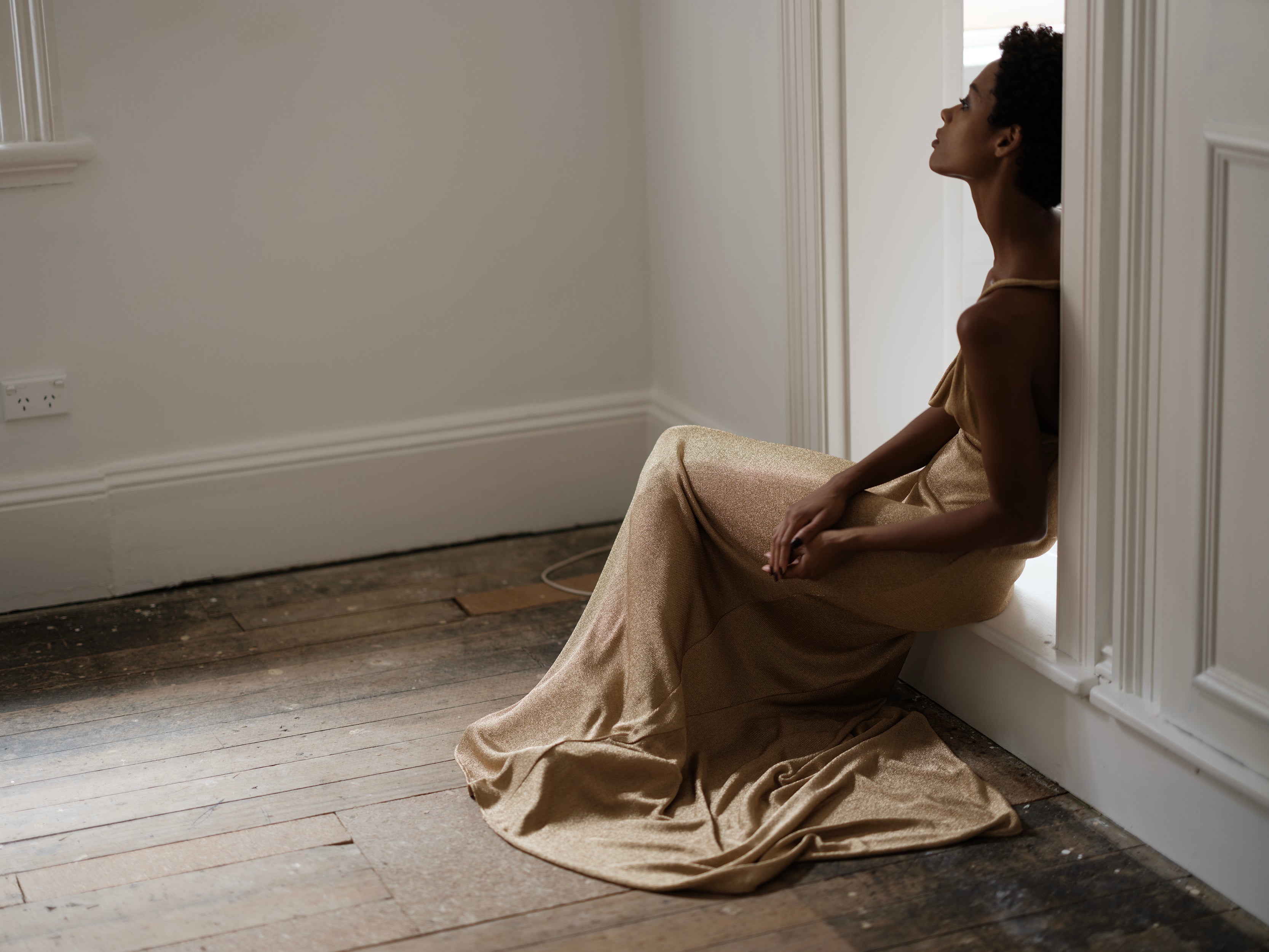 Woman in a Common Hours metallic gold draped dress sitting on a wooden floor in a minimalistic room.