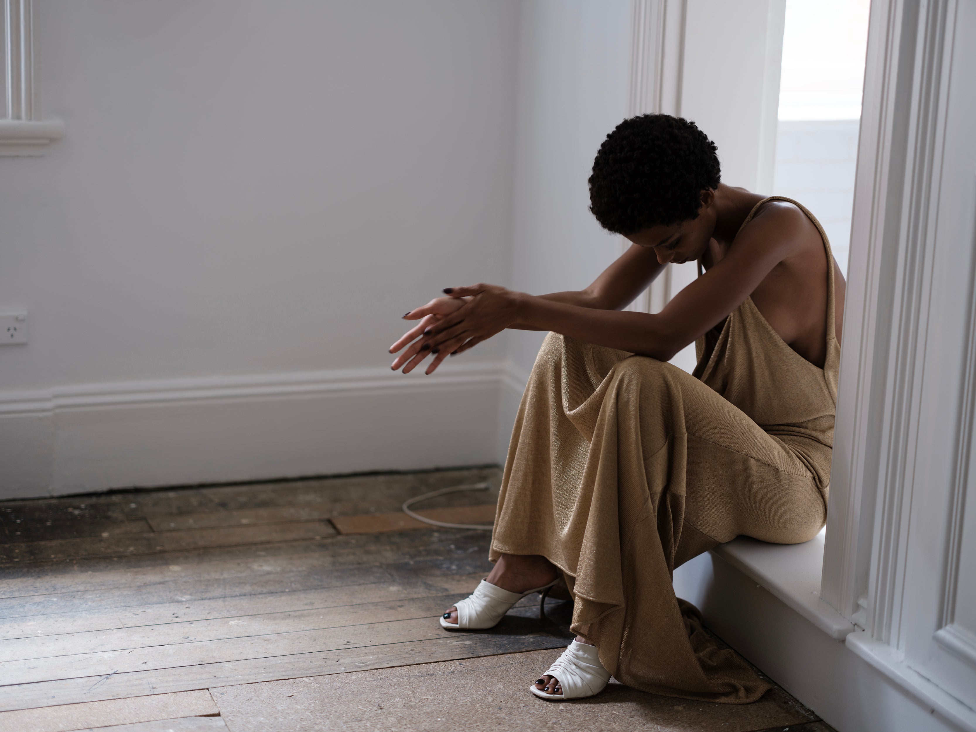 Model wearing a draped Common Hours gold metallic dress sitting on a door frame ledge in a minimalistic room with wooden floor and white walls.