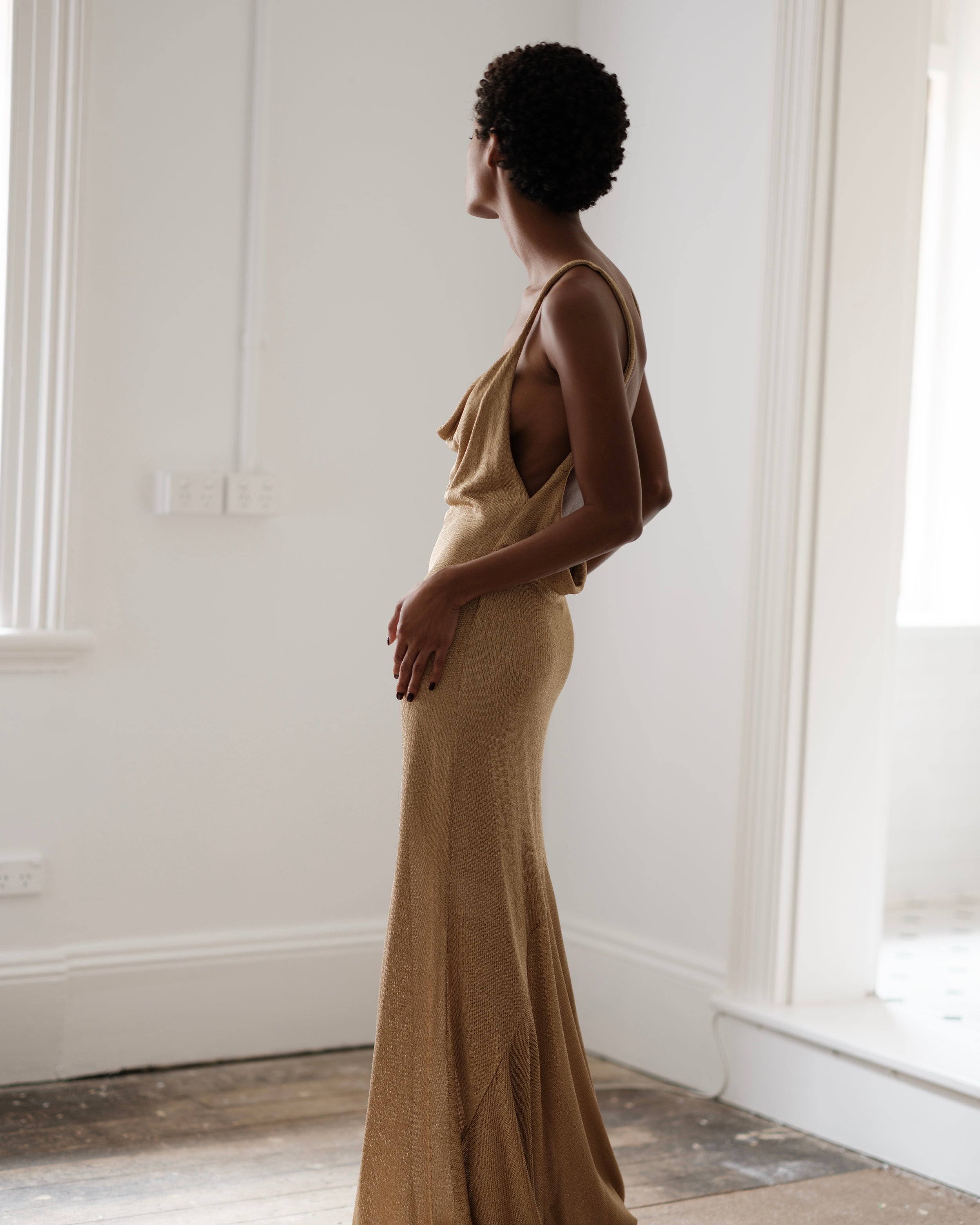 Woman in a Common Hours gold draped, backless dress standing in a room with white walls and wooden floor.