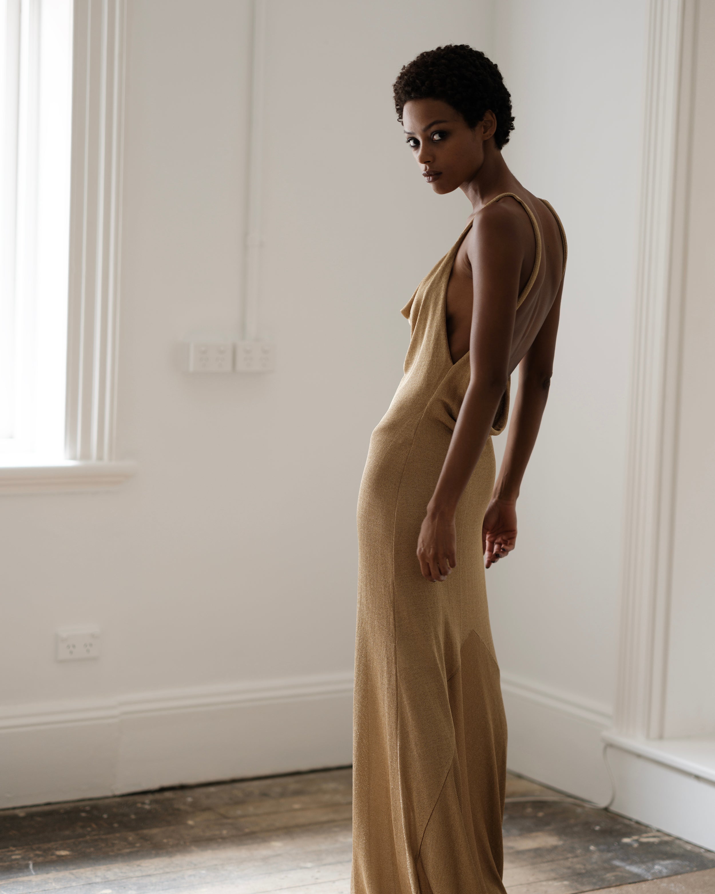 Woman in a gold Common Hours backless dress standing in a room with white walls and wooden floor.