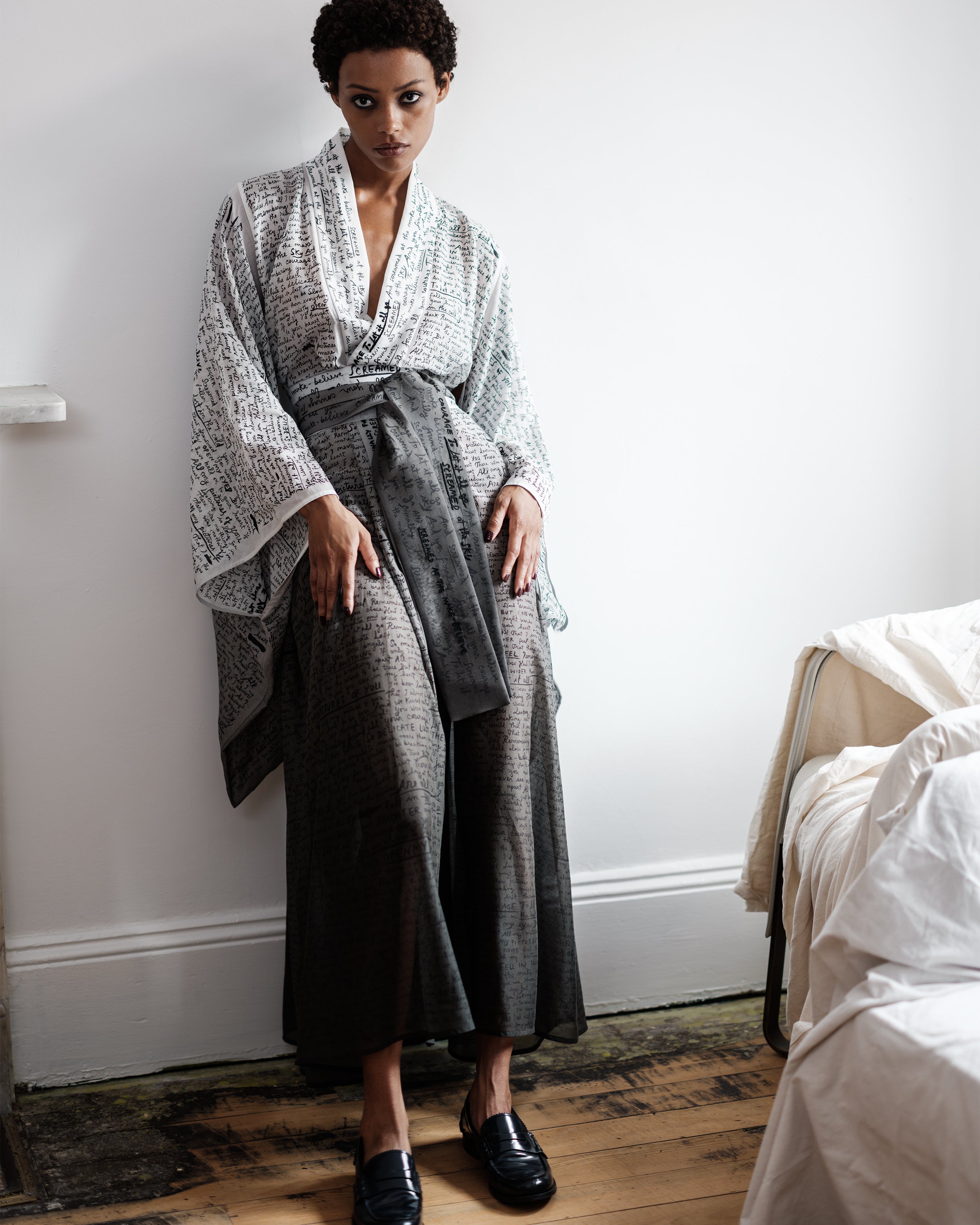 Woman wearing a Common Hours wrap dress with printed and hand-embroidered words, standing in a bedroom.
