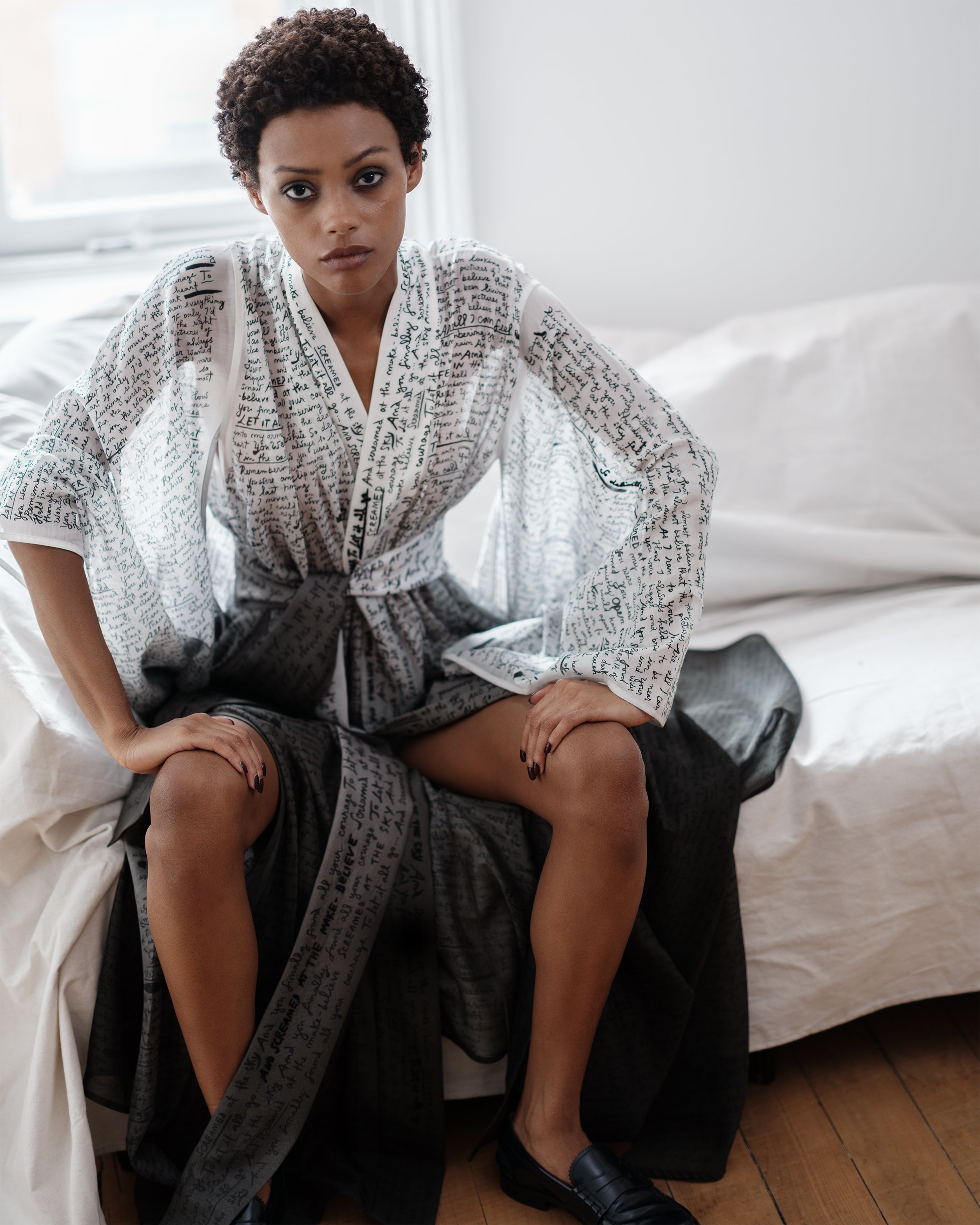 Woman wearing a Common Hours wrap dress with printed and hand-embroidered lyrics sitting on a bed in a bedroom setting.